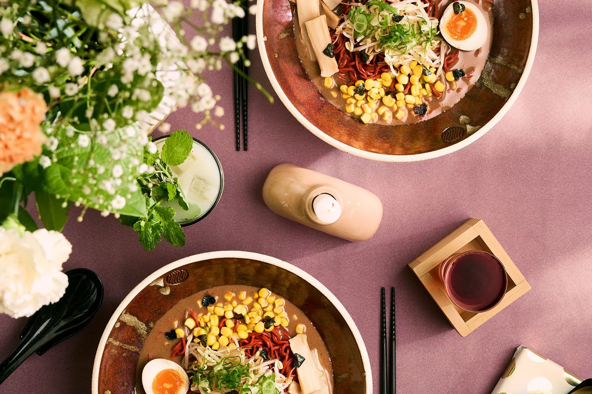 Two bowls of ramen with corn, half-boiled eggs, green onions, and bamboo shoots on a purple tablecloth with chopsticks, drinks, flowers, and a candle.