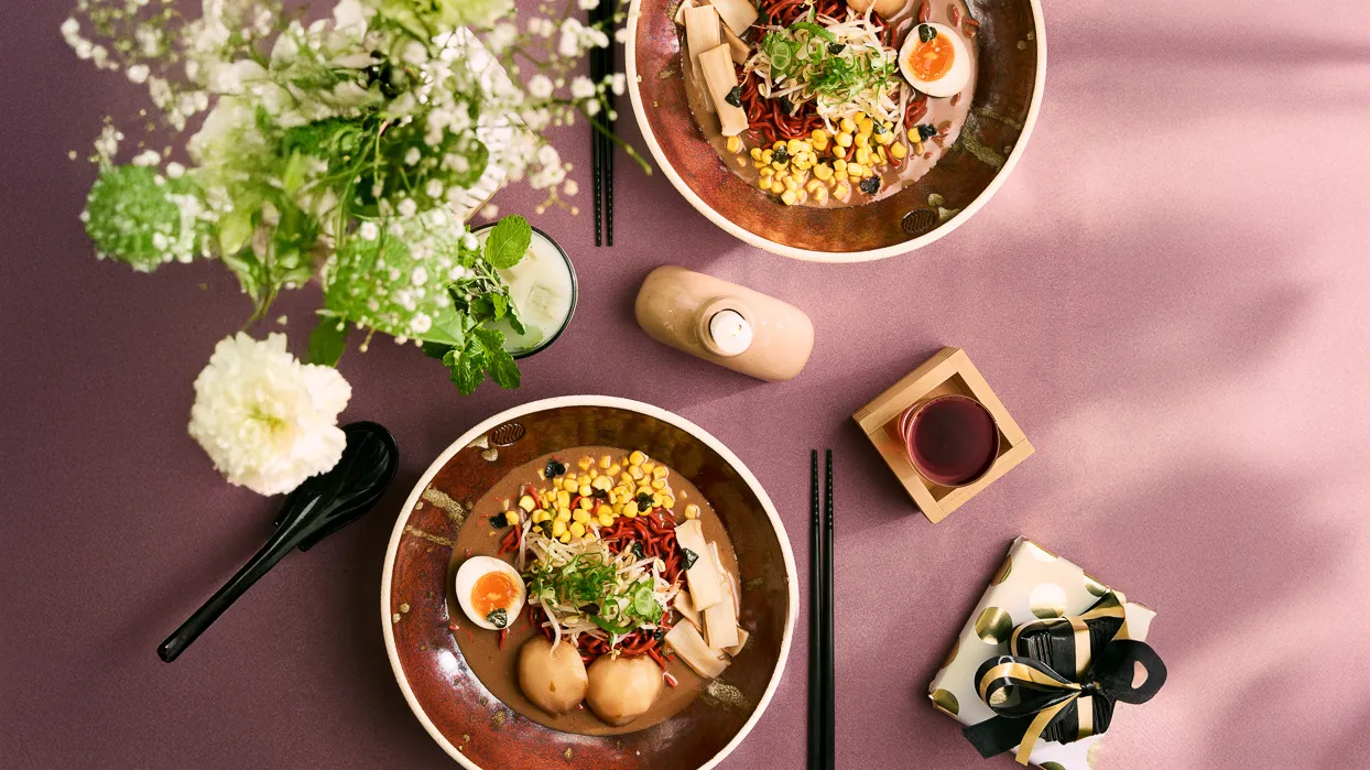 Top-down view of two bowls of ramen with corn, soft-boiled egg, bamboo shoots, and green onions on a pink table with chopsticks, a black spoon, a cocktail with mint, a small red drink, greenery, and a wrapped gift box.