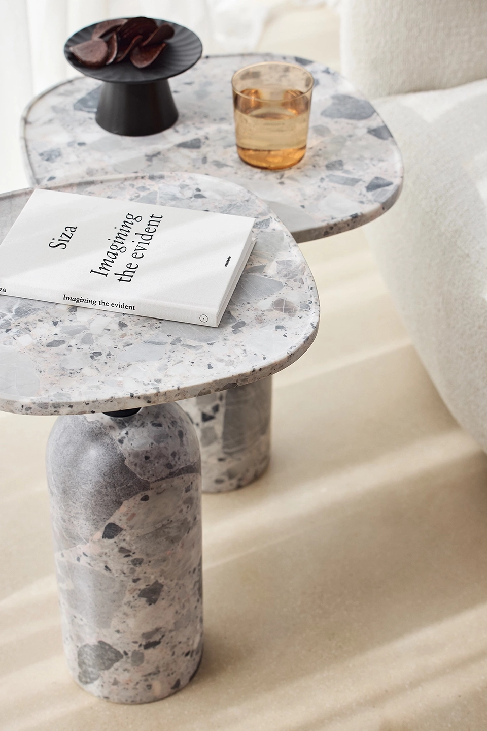 Two terrazzo side tables with rounded tops; the nearer table holds a white book titled 'Imagining the evident' and the farther table has a small amber glass and a black pedestal dish with dark chocolate pieces.