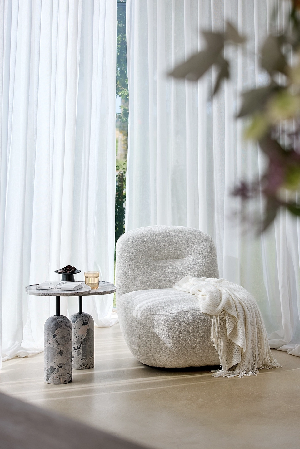 Modern white bouclé armchair with a textured cream throw draped over it next to a round marble-topped side table holding a book, glass, and decorative bowl in front of sheer white curtains.
