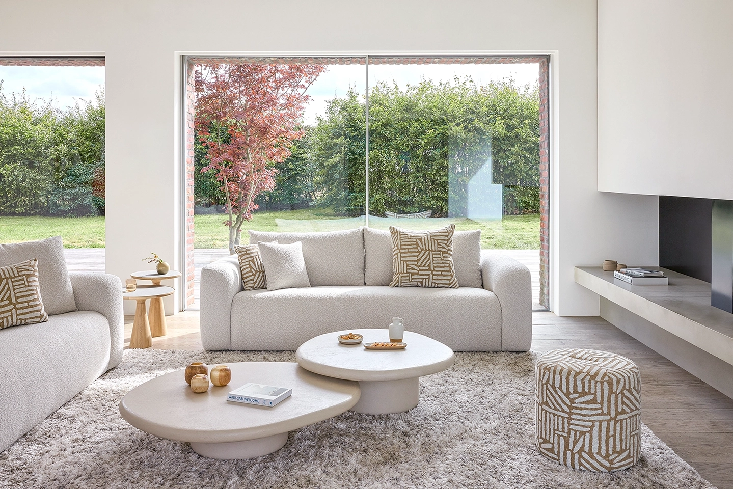 Bright modern living room with cream textured sofas, patterned cushions, circular coffee tables, and large windows showing a garden.