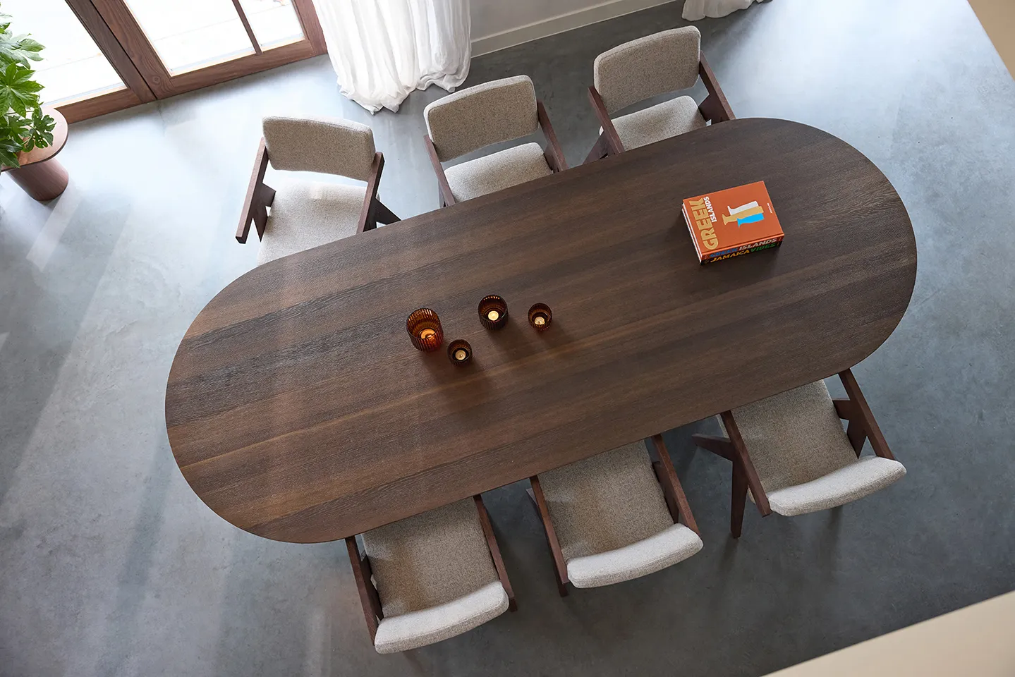 Top view of a dark wooden oval dining table with six beige cushioned chairs, three orange candle holders, and a travel book on a polished concrete floor near a glass door.