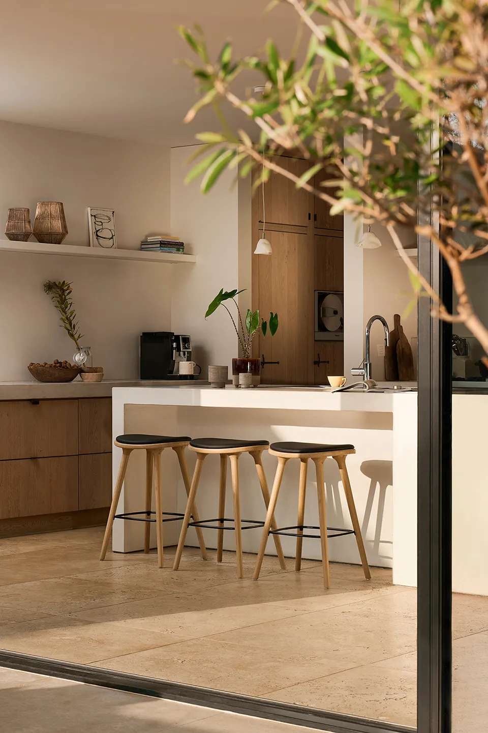 Sunlit modern kitchen with three wooden stools at a white island countertop, wooden cabinets, a coffee machine, and decorative plants.