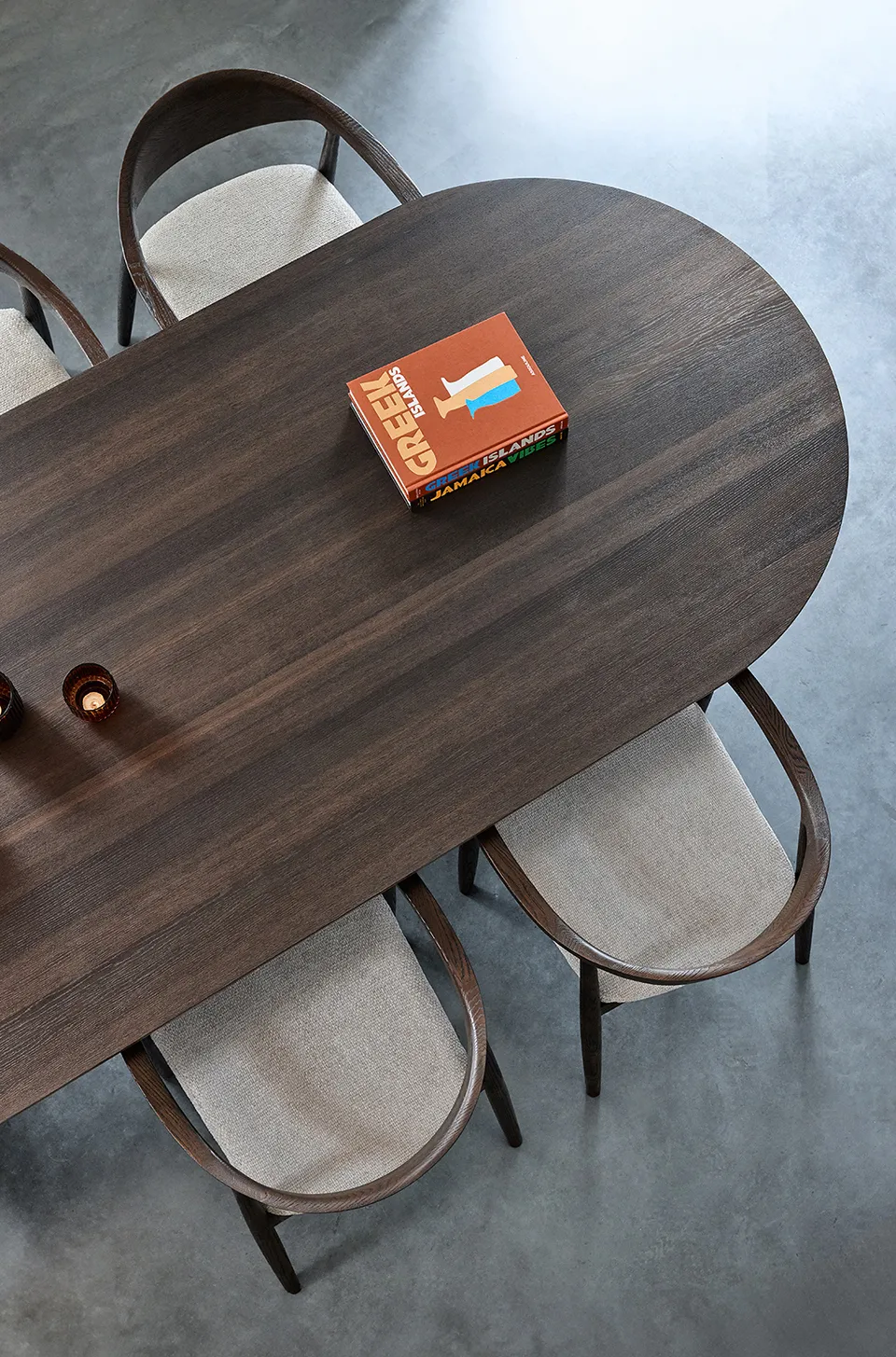 Top view of a dark wooden oval table with three cushioned chairs and a book titled 'Greek Islands' on it.