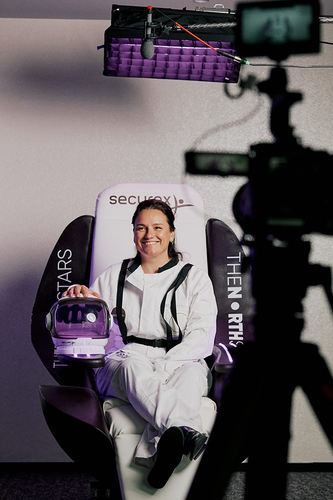 Smiling woman in a white spacesuit sitting on a chair labeled THENORTHSTARS, holding a space helmet, with recording equipment in the foreground.