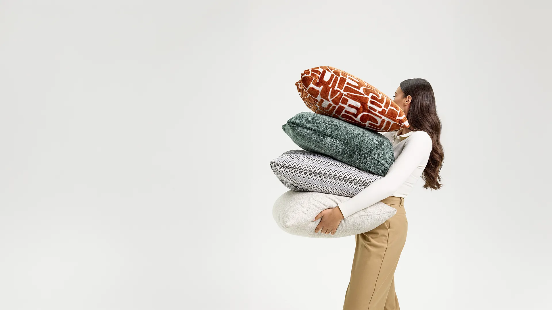 Woman in beige pants and white top carrying a stack of four decorative pillows with different patterns.