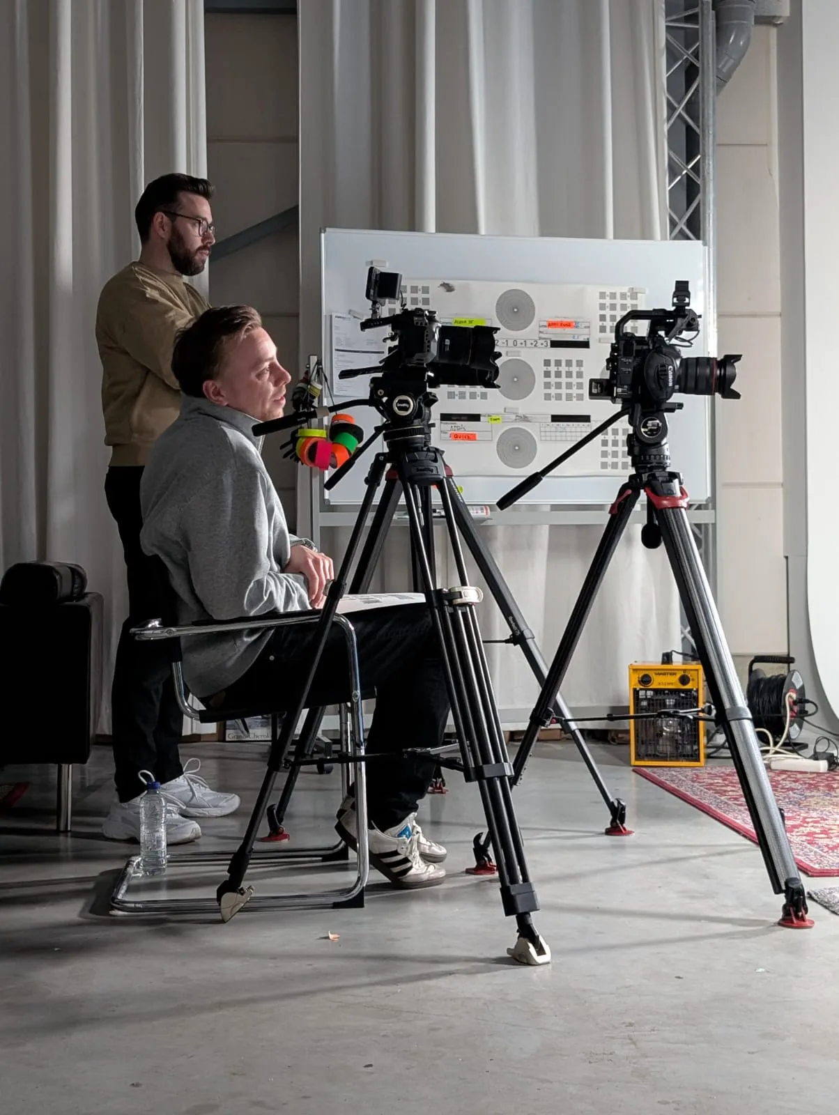 Two men in a studio setting; one seated behind a camera on a tripod, the other standing nearby with a whiteboard and equipment in the background.
