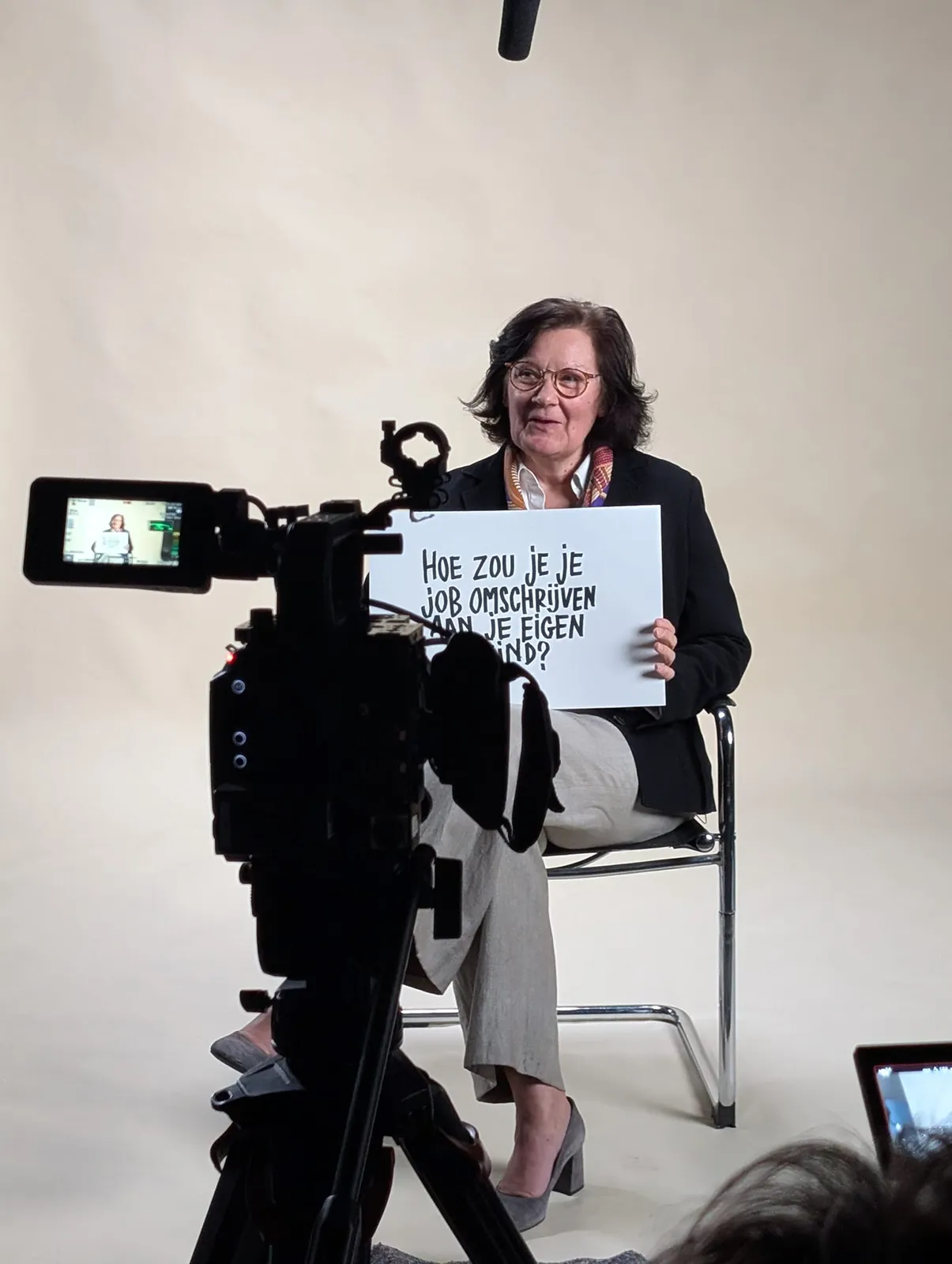 Woman sitting on a chair in a studio holding a sign with Dutch text while being recorded on camera.