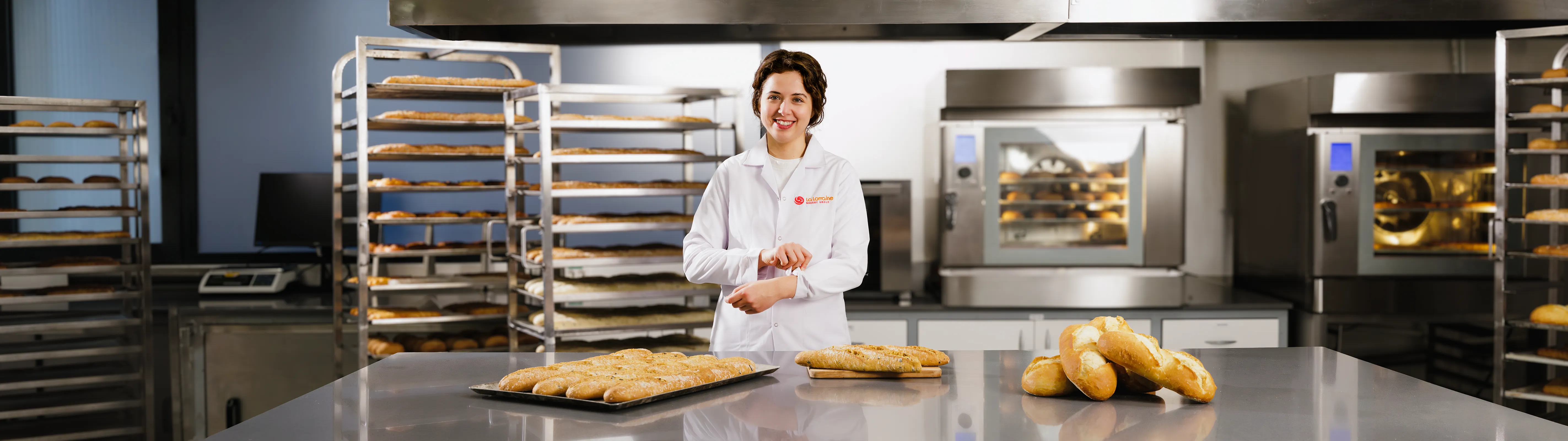 Smiling baker in white coat standing behind stainless steel table with freshly baked bread in a commercial bakery kitchen.