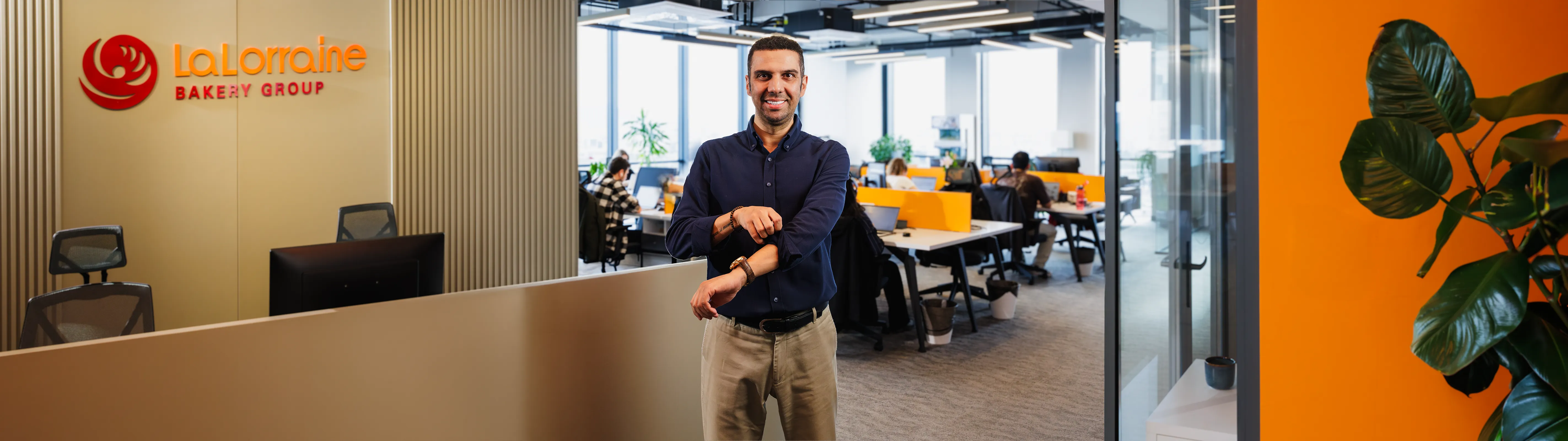 Smiling man standing in modern office reception area with La Lorraine Bakery Group logo, and employees working at desks in background.