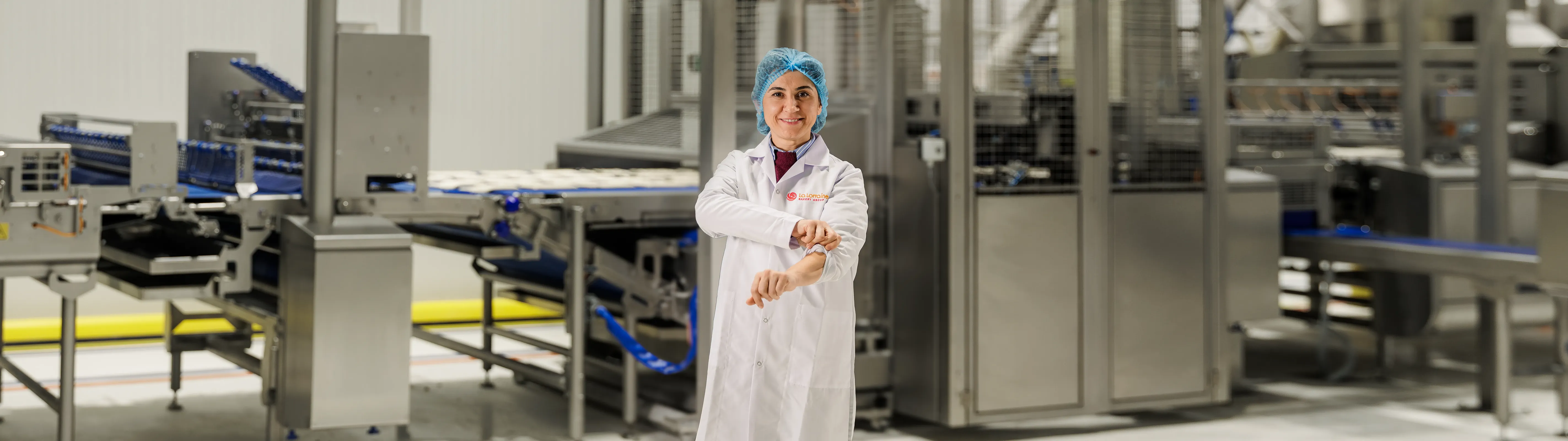 Smiling factory worker in a white coat and blue hairnet adjusting her sleeve in a food processing facility.