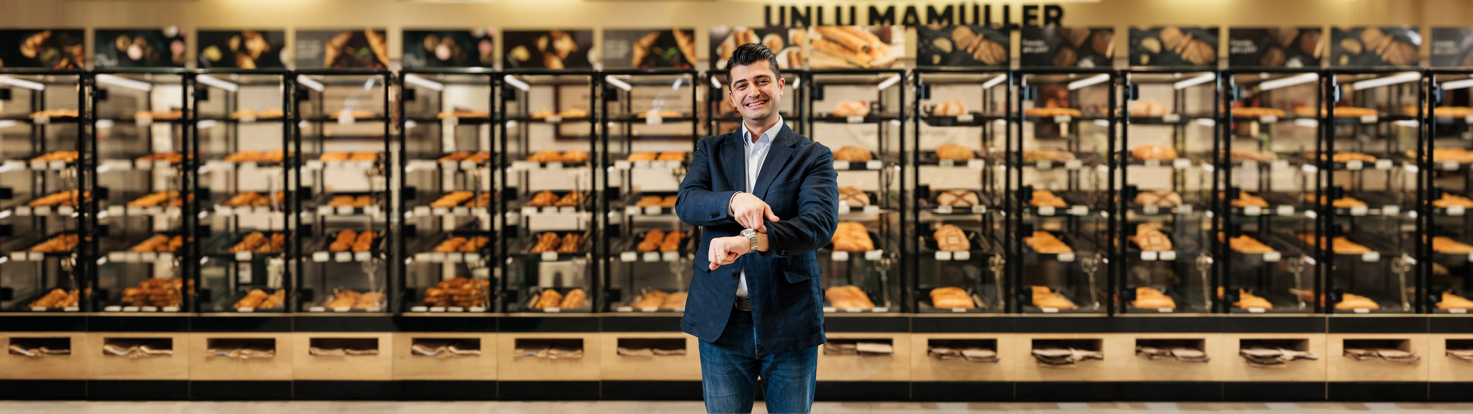 Smiling man in a dark blazer pointing at his wristwatch in front of glass bakery display cases filled with various breads and pastries.