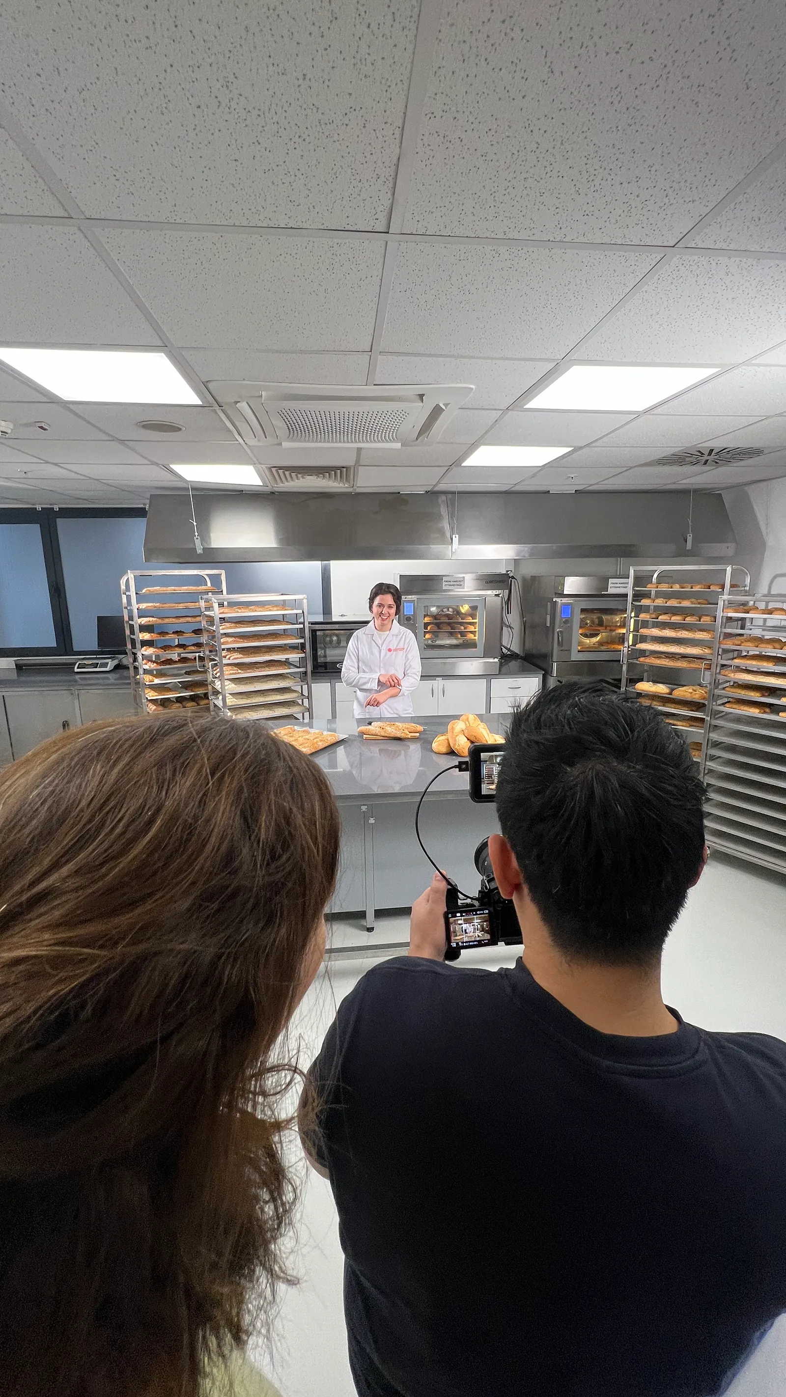 A baker in white uniform standing behind a table with bread loaves in a commercial kitchen while two people film with a camera.