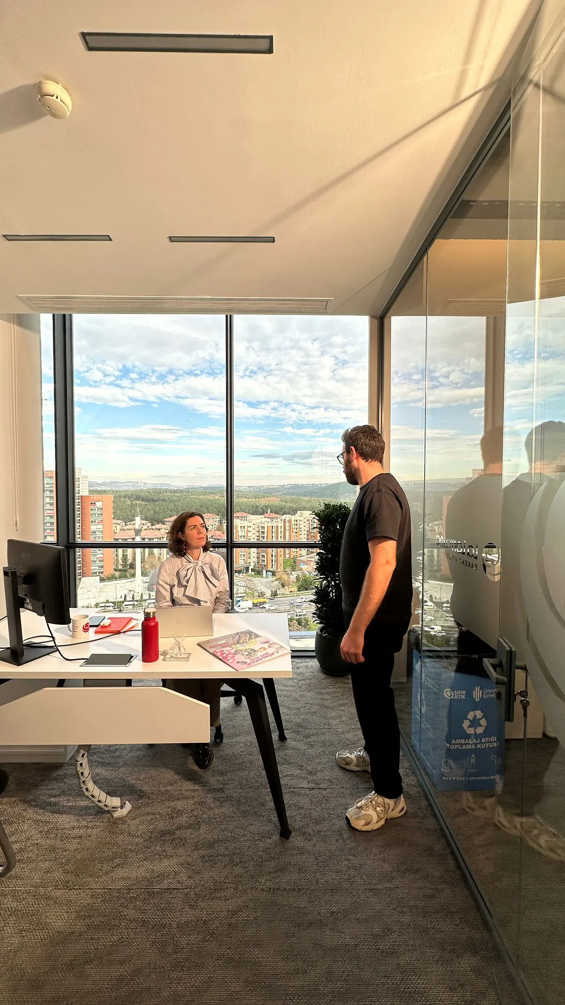 Woman sitting at a desk with a laptop talking to a standing man in a modern office with large windows showing a cityscape.