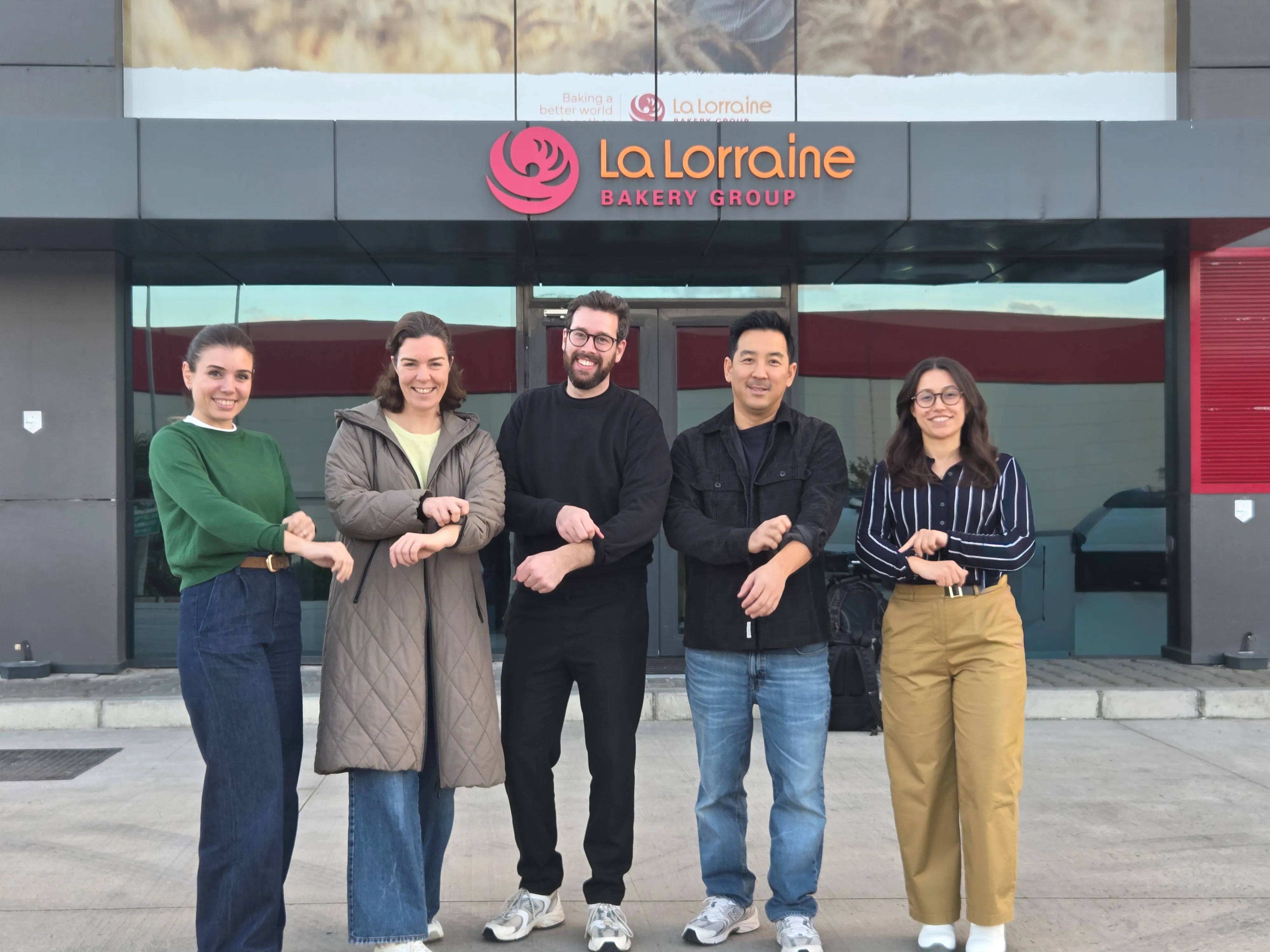 Five people smiling and standing outside La Lorraine Bakery Group building, showing a wrist gesture.