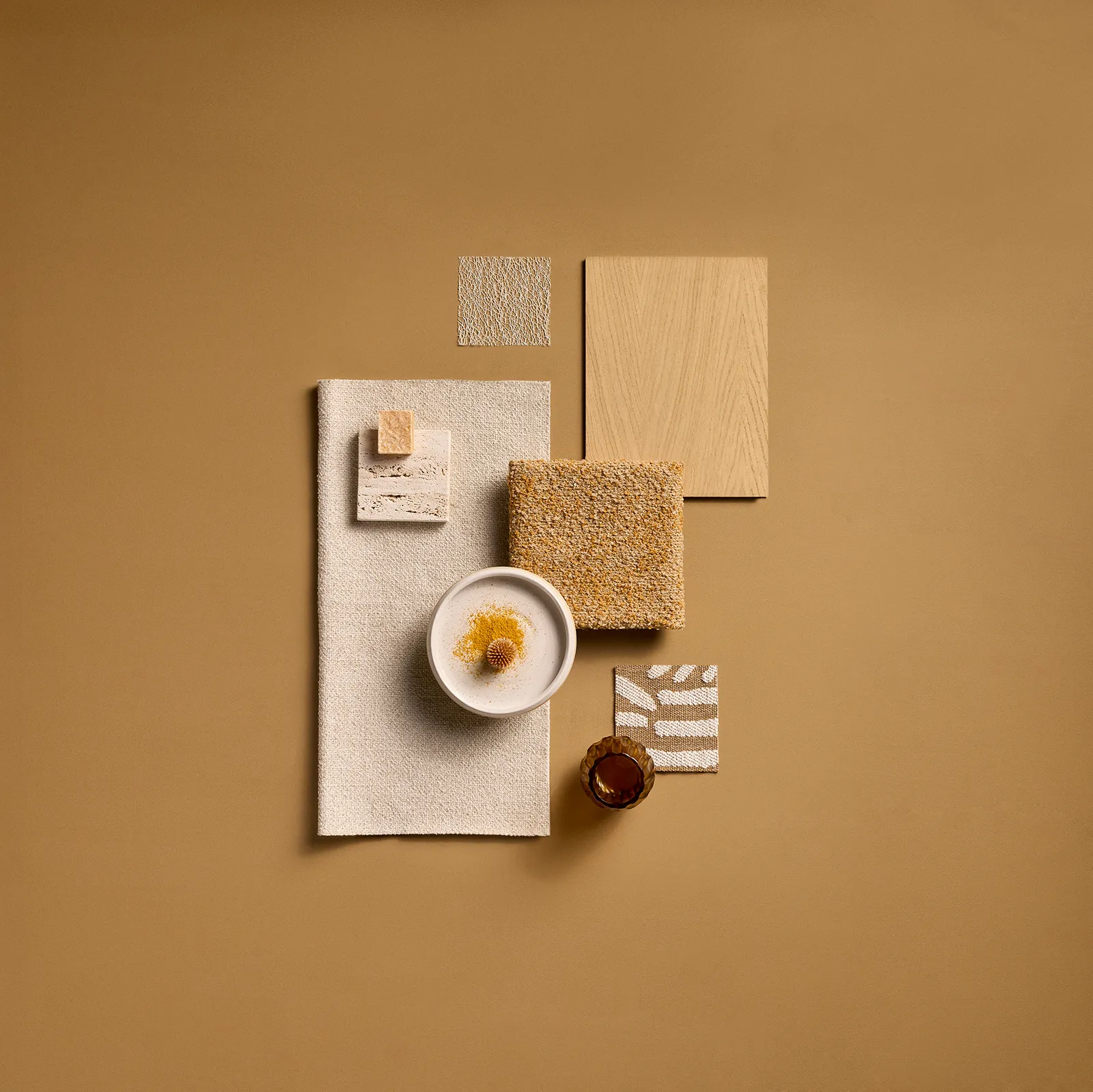 Flat lay of beige and brown textured materials and objects arranged on a tan background, including fabric swatches, wood panel, a small bowl with a dried flower, and a brown glass.