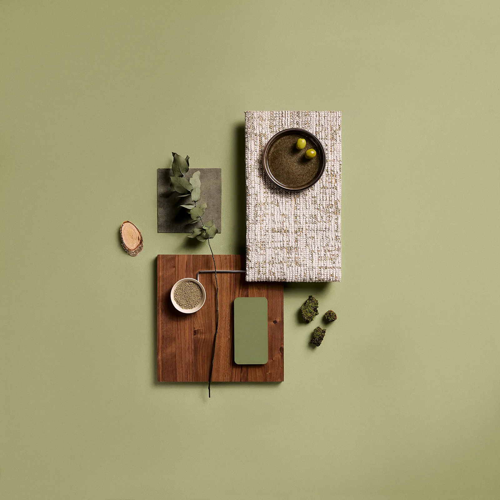 Flat lay of natural and green elements including a wooden board, a textured fabric swatch, a small dish with green powder, a twig with green leaves, green paint sample, moss pieces, and a bowl with two green olives on a light olive green background.