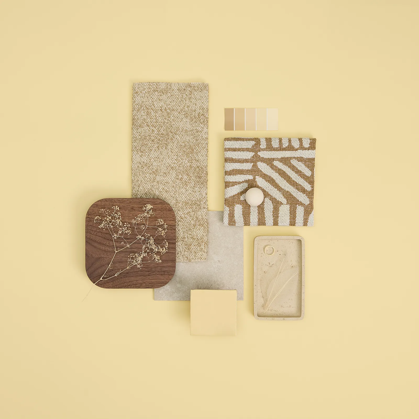Flat lay of neutral-tone design materials including wood with dried flowers, textured fabrics, paint swatches, and ceramic tray with dried leaves on pale yellow background.