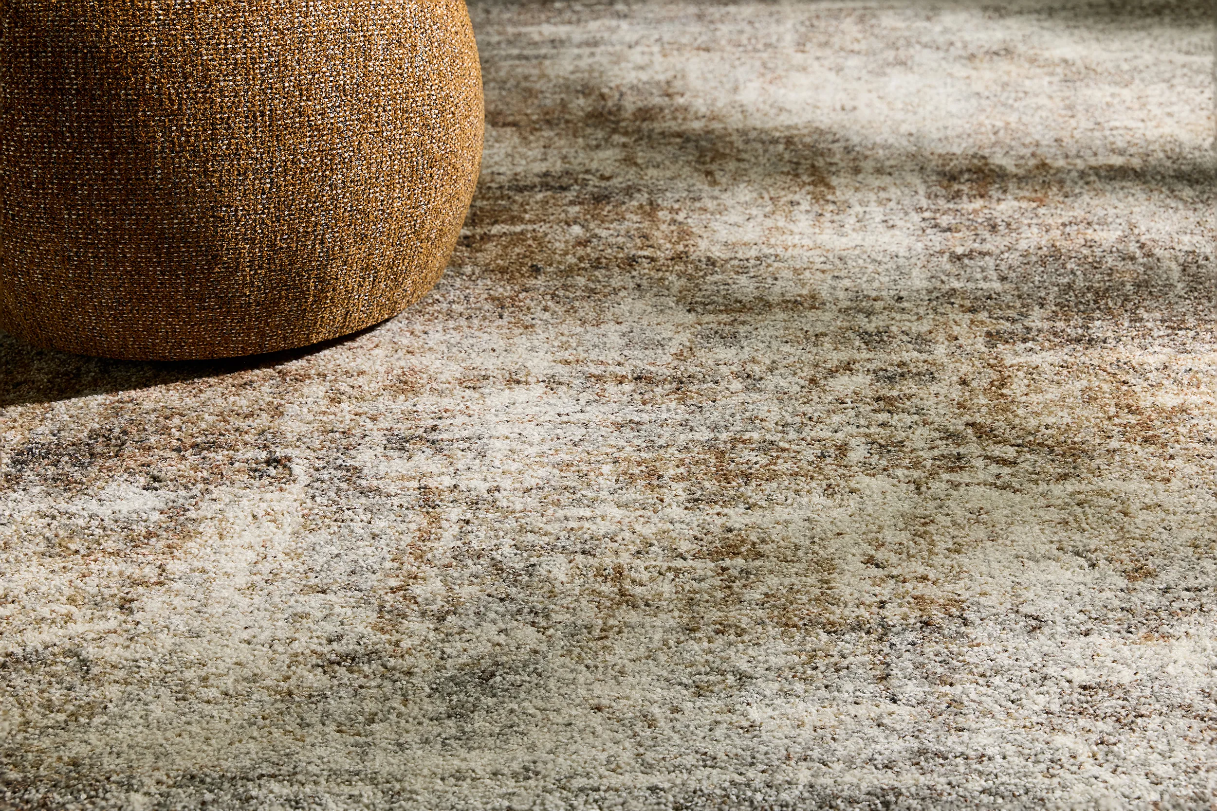 Close-up of a textured brown woven pouf on a carpet with a mottled pattern in shades of beige, brown, and gray.