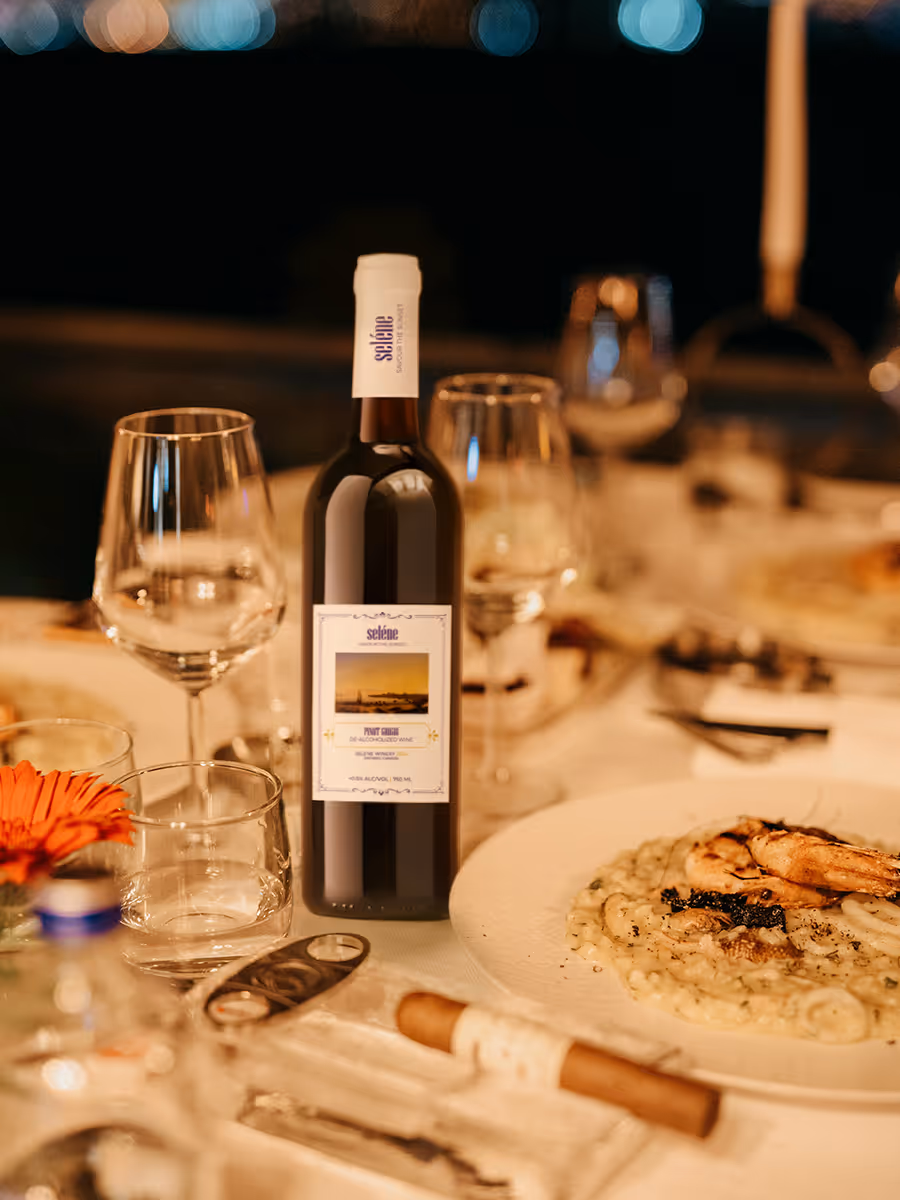 Dining table set with a bottle of Selene Pinot Grigio wine, empty wine glasses, a plate of risotto with shrimp, and a cigar in a glass ashtray.