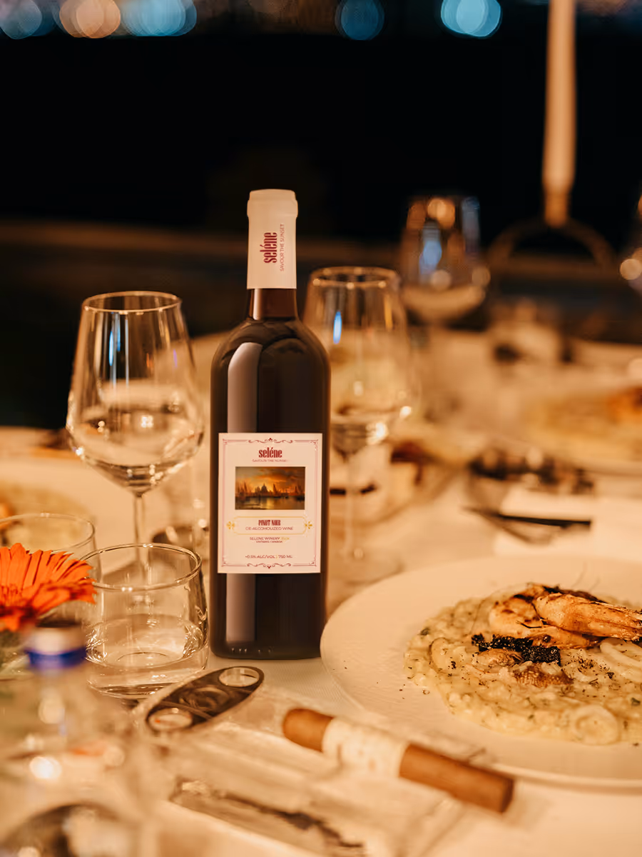 Bottle of Selene Pinot Noir wine on a dinner table with glasses, a flower, cigar, and a plate of food under warm lighting.