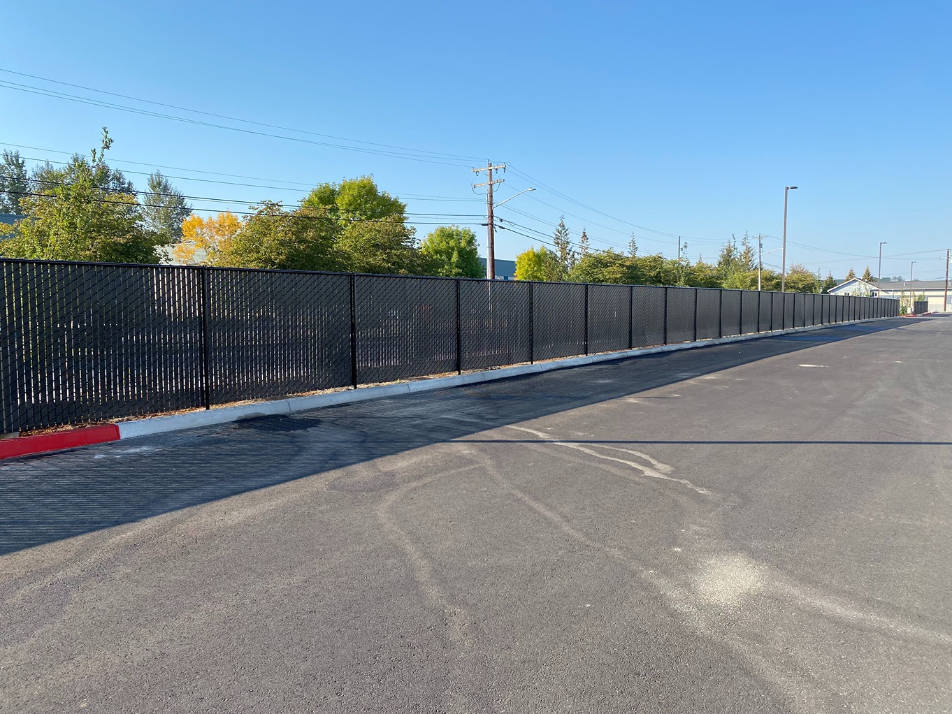 A long, black chain-link fence with privacy slats lines the edge of a freshly paved asphalt parking 