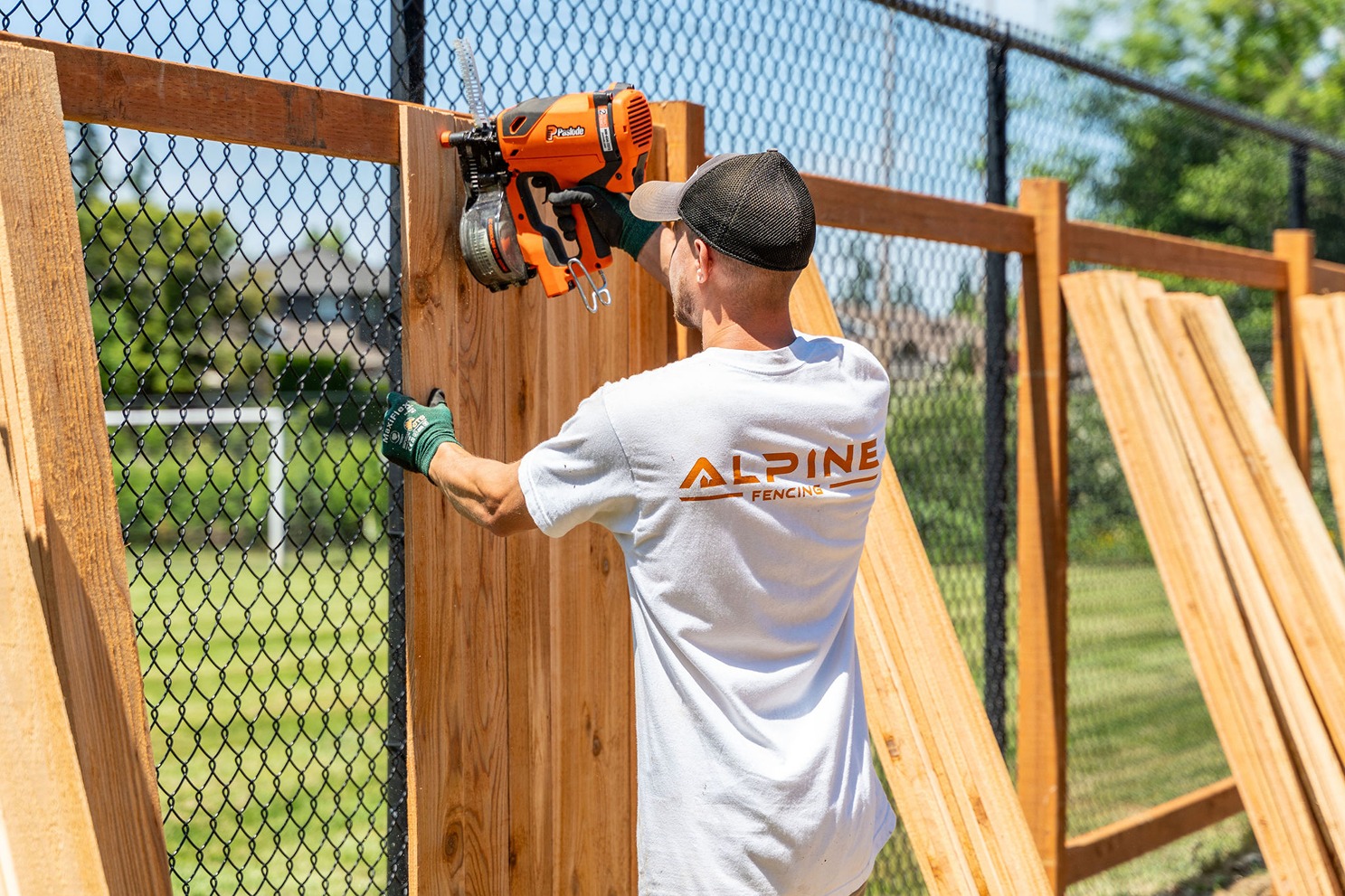worker wearing a white "Alpine Fencing" t-shirt and green gloves installs vertical wooden panels onto a black chain-link fence using a nail gun