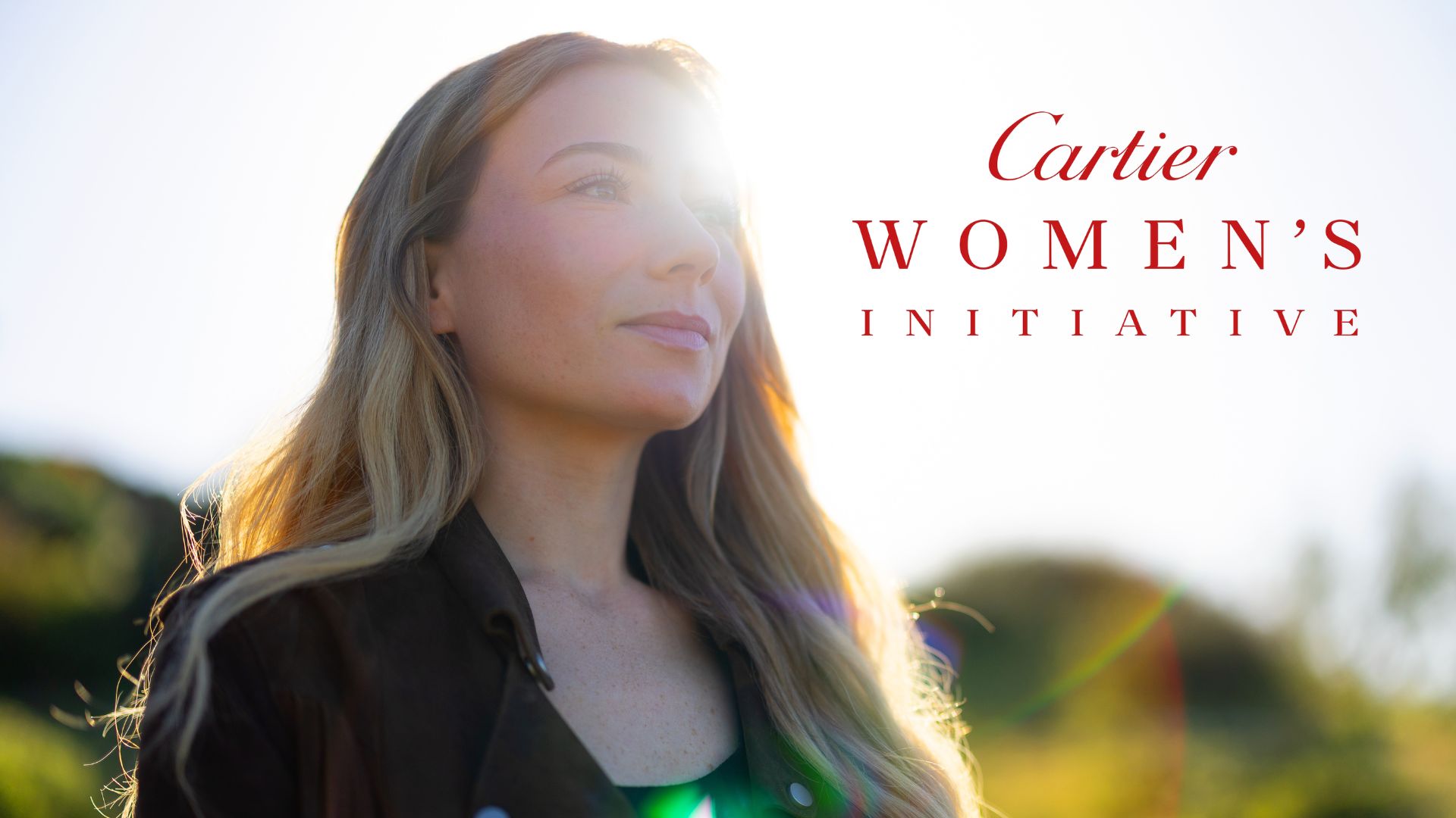 A woman with long light-brown hair stands outdoors in warm sunlight, looking upward with a calm, confident expression. Lens flare creates a soft, glowing effect across the image. On the right side, red text reads “Cartier Women’s Initiative.”