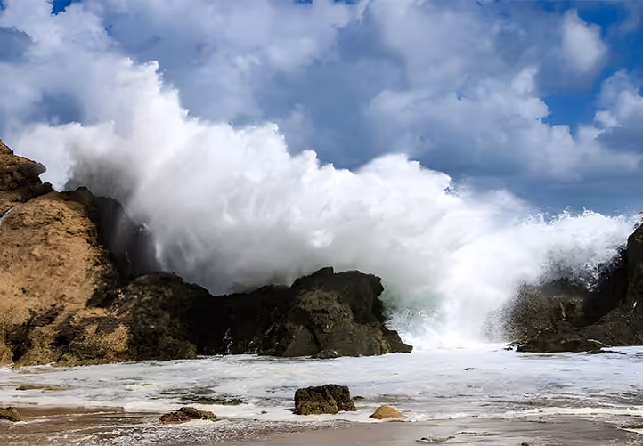 A powerful ocean wave crashes against rocky cliffs, creating a dramatic spray of white water that towers high into the air. The scene shows a sandy beach with scattered rocks under a blue sky filled with white and gray clouds.