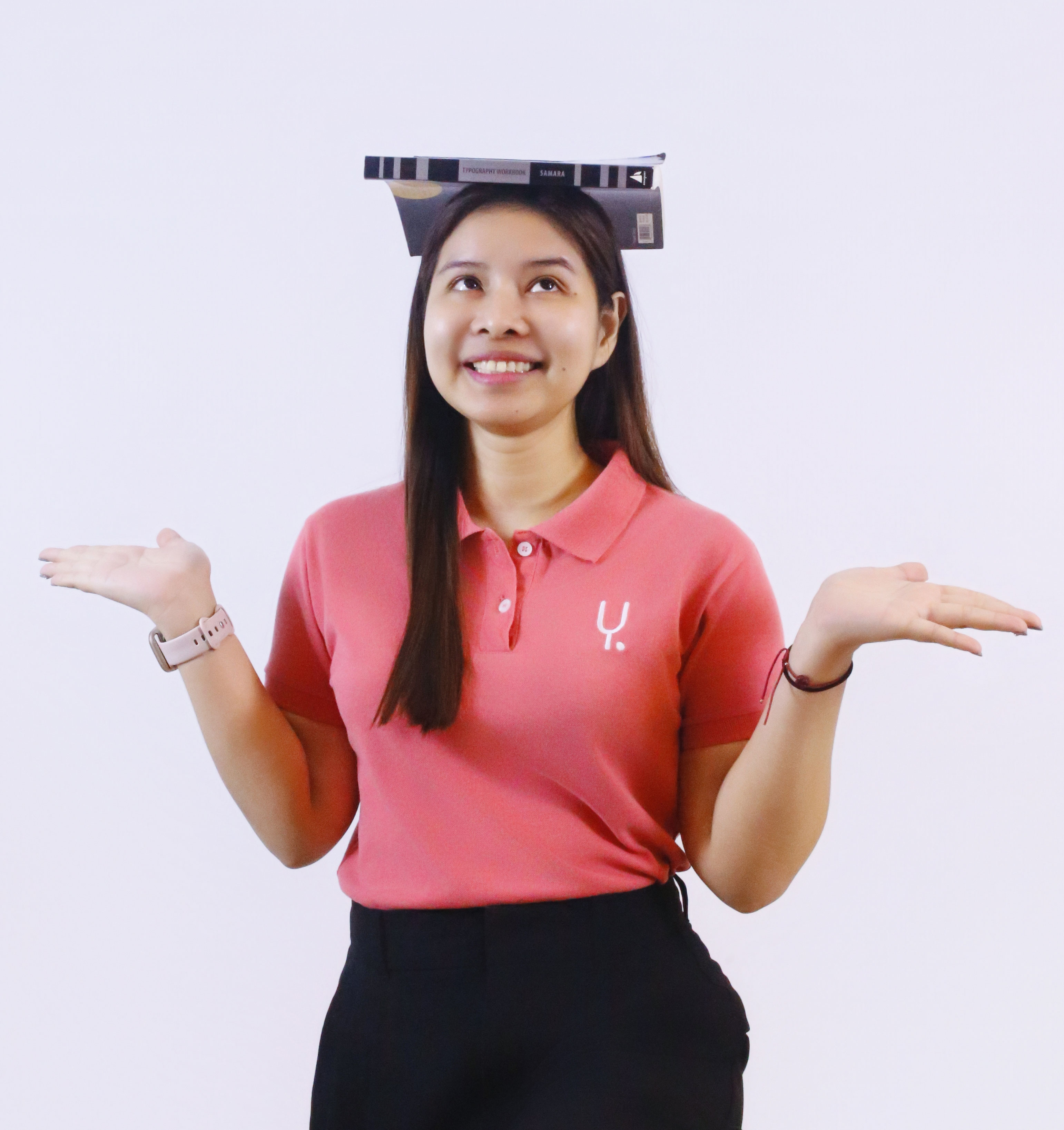 A smiling young woman with long dark hair balances a small stack of books on her head while holding both hands out to her sides in a playful balancing gesture. She's wearing a coral pink polo shirt and black skirt, looking up and to the side with a cheerful expression against a light purple background.