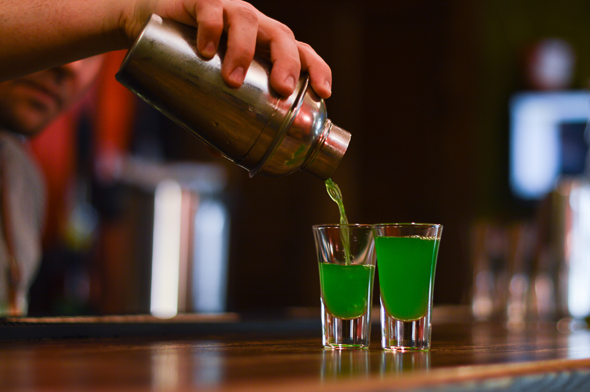 A hand pouring a bright green liquid from a metal cocktail shaker into two shot glasses on a wooden bar counter.