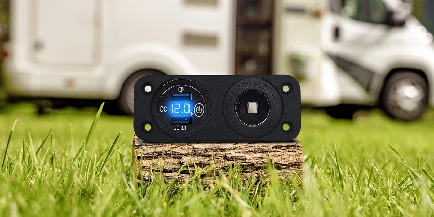 USB-laadpaneel met digitale voltmeter (12 V, QC 3.0) staat op een houtblok in het gras; gefotografeerd met een camper op de achtergrond