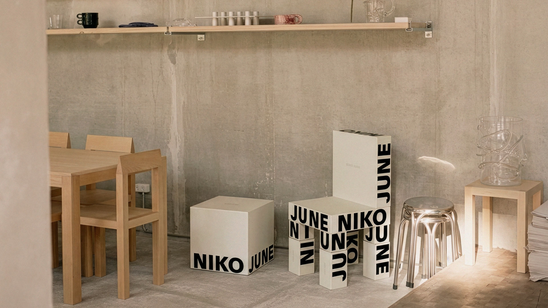 Sculptural Boxes in Studio Interior Interior scene featuring sculptural branded boxes for NIKO JUNE stacked playfully in a concrete space with minimalist furniture and art objects. The packaging, designed by N96 — a Copenhagen-based brand studio — uses large-scale typography and spatial composition to reflect the brand’s experimental, gallery-inspired aesthetic.