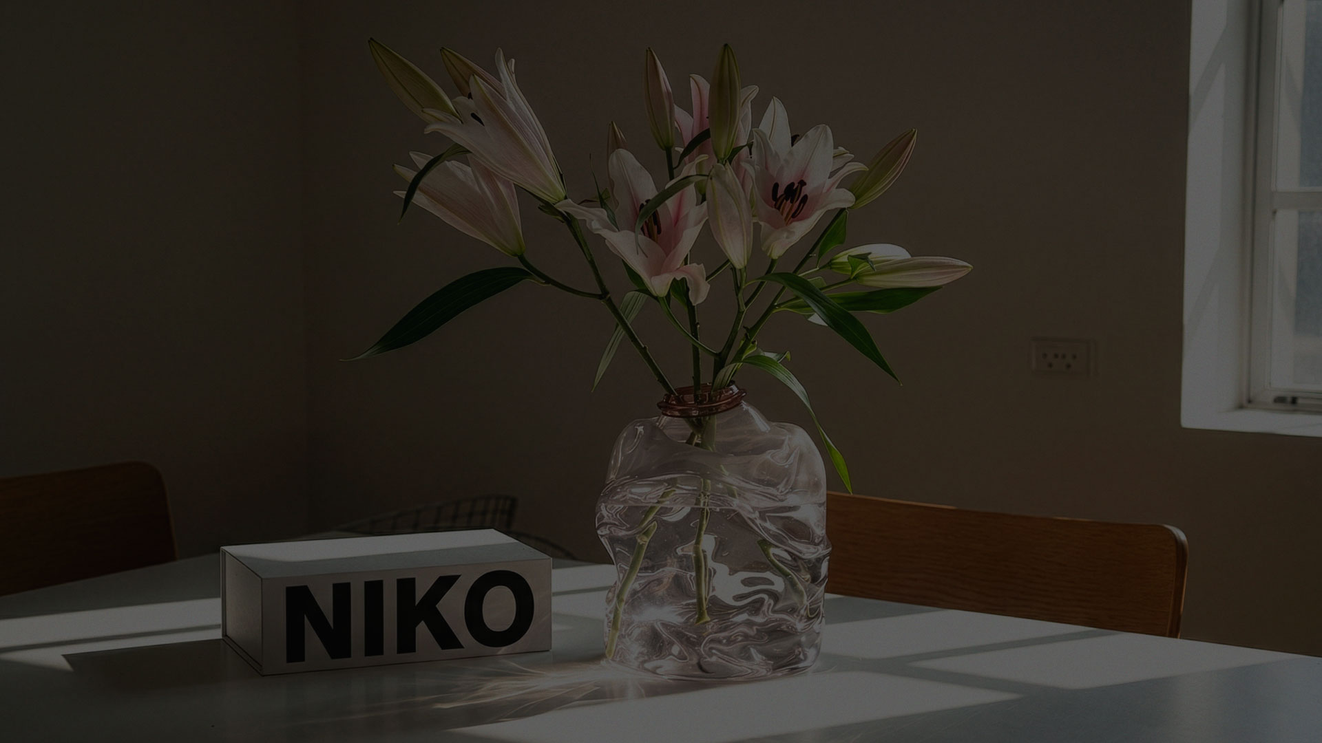 A sunlit table scene with a soft pink glass vase holding blooming lilies, next to a rectangular white box marked “NIKO.” The minimal setting and play of natural light evoke quiet beauty and thoughtful placement. Created by N96, this brand expression captures the poetic simplicity and object-as-sculpture ethos central to Niko June’s universe.