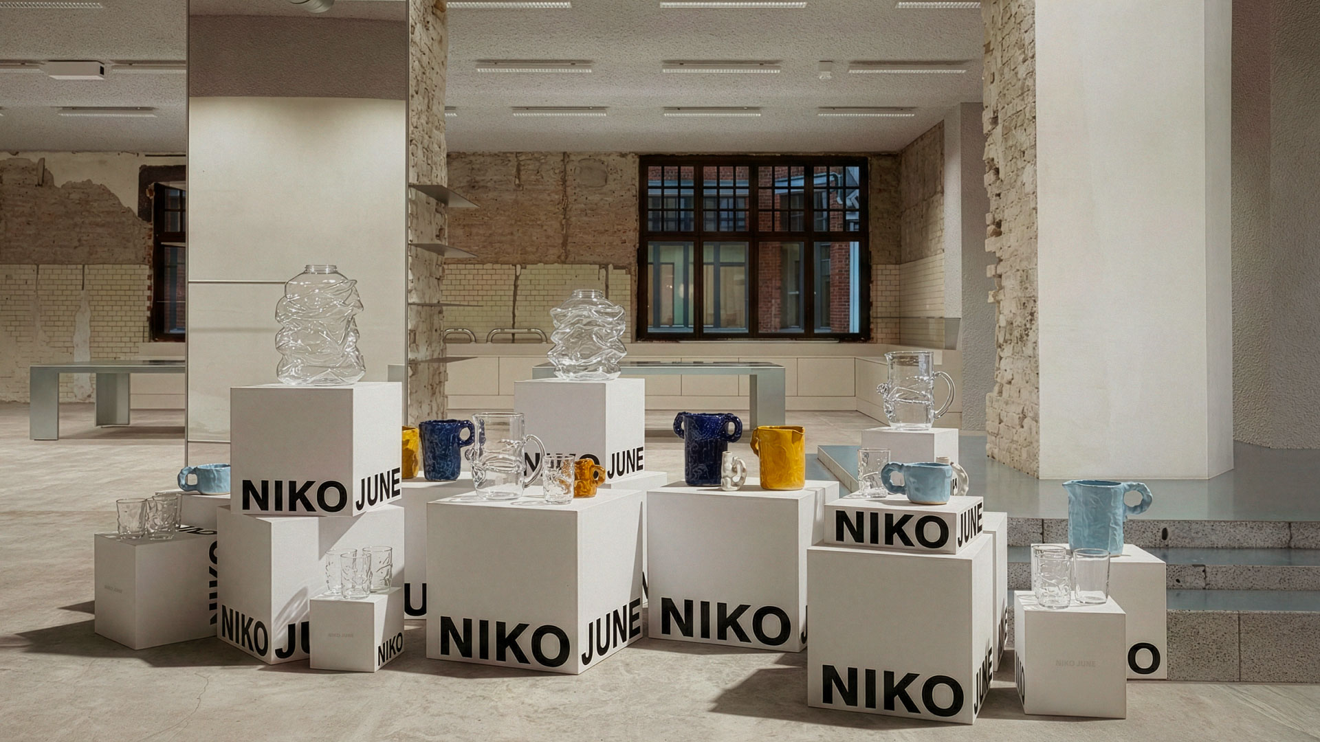 A minimalist showroom features a display of stacked white boxes branded with “NIKO JUNE” in bold black typography. Atop the boxes are various artistic glassware pieces in clear, amber, blue, and yellow tones — including crumpled vases, mugs with textured handles, and tumblers. The backdrop includes a raw, industrial space with exposed brick walls, a mirror pillar, and large windows, creating a refined yet unfinished aesthetic.