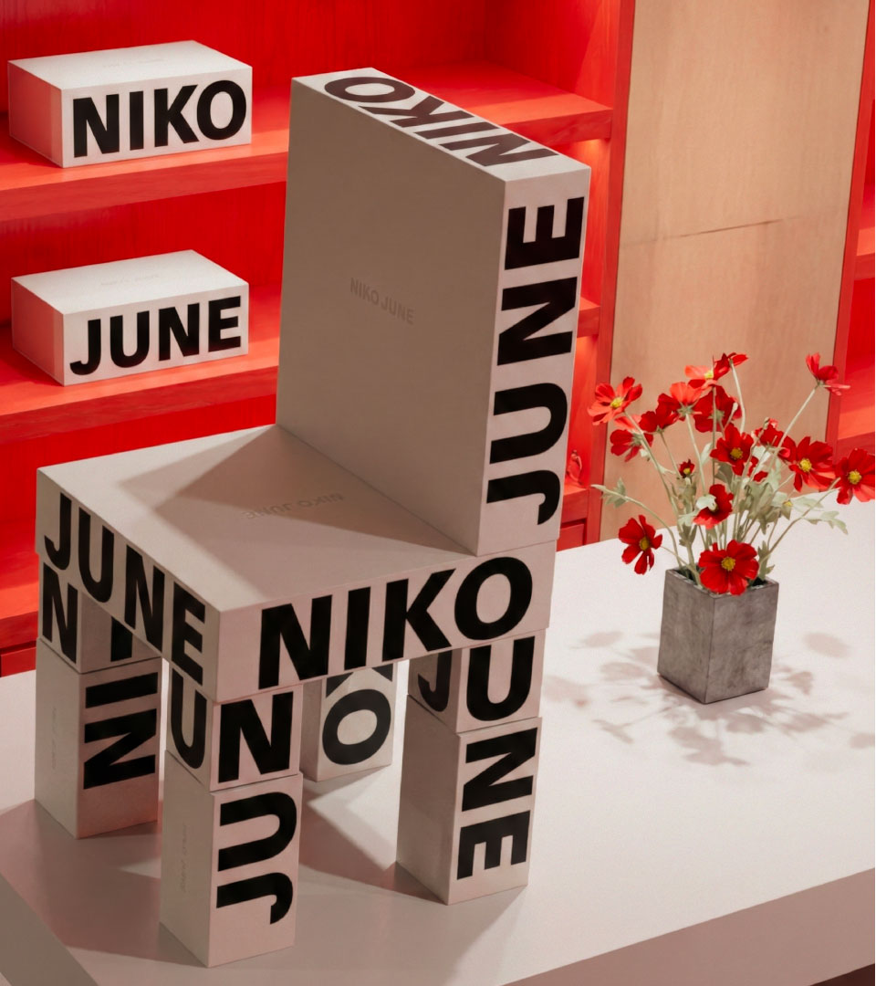 A striking branded chair constructed entirely from modular packaging blocks designed for NIKO JUNE, displayed in a vivid red retail environment. The boxes feature bold black typography with the brand’s name, forming both the structure and aesthetic of the furniture. A minimal white surface and a vase of red flowers create visual balance and softness. Designed by N96, a Copenhagen-based creative studio specializing in consumer brand identity and design, this project exemplifies innovative brand activation, spatial branding, and physical storytelling for lifestyle and design-forward brands.