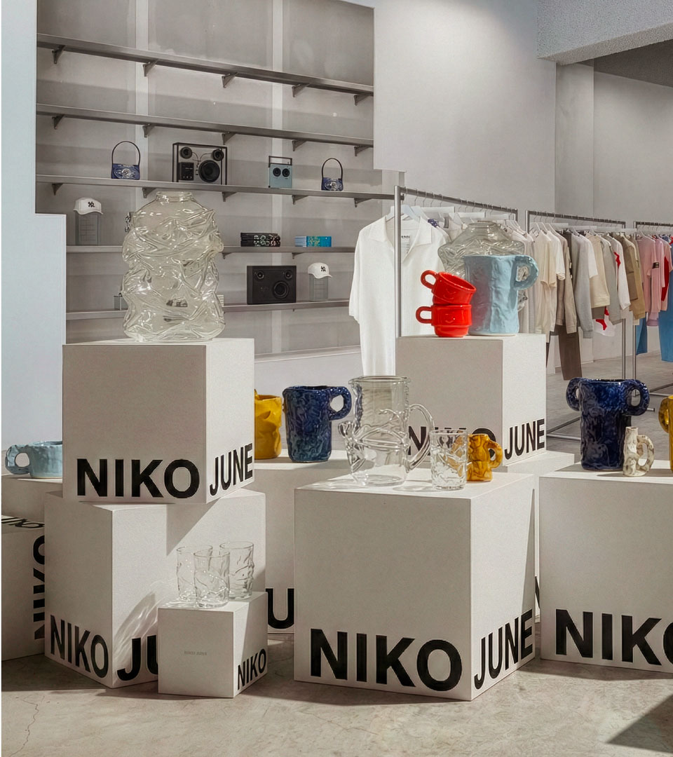Branded retail experience design for NIKO JUNE, featuring minimalist white display cubes with bold black typography spelling out the brand name. The cubes showcase an eclectic mix of sculptural glass objects and ceramic pieces inside a concept store environment. Created by N96, a Copenhagen-based creative consumer brand studio specialising in strategic branding, identity, and cultural resonance for design-forward and challenger brands.