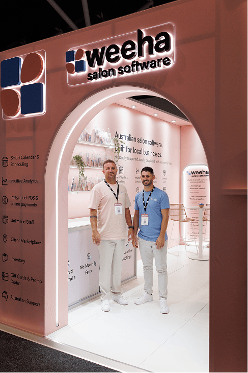Weeha founders Jace and George standing in the arched entrance of the pink Weeha salon software stand