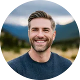 Smiling man in a casual sweater, standing outdoors with a mountain backdrop and cloudy sky.