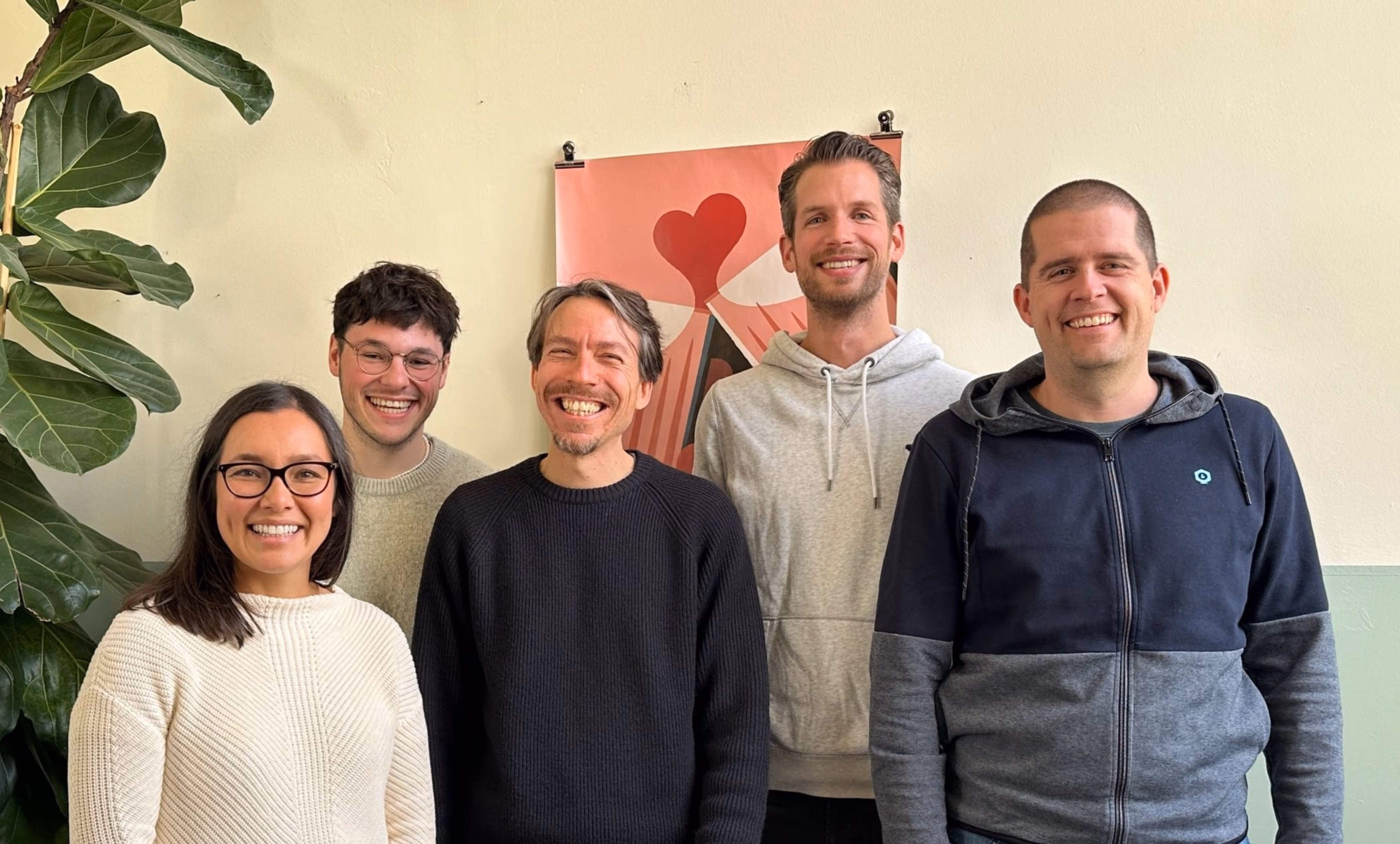 Fünf lächelnde Personen stehen vor einer Wand mit einem Poster, daneben eine große grüne Pflanze.