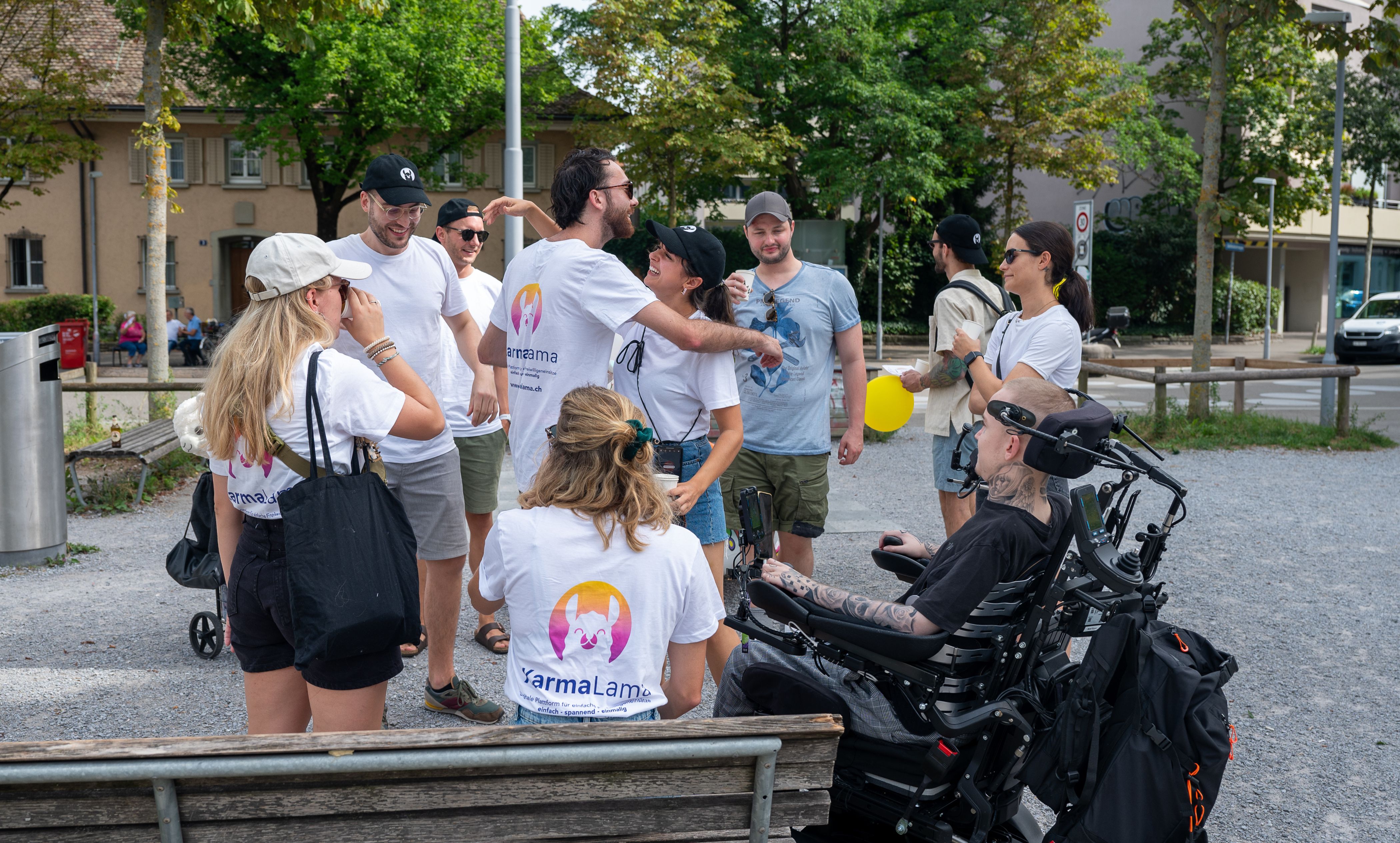 Gruppe von Menschen in weißem Shirt mit YarmaLama-Logo, die im Freien lachen, sich umarmen und unterhalten.