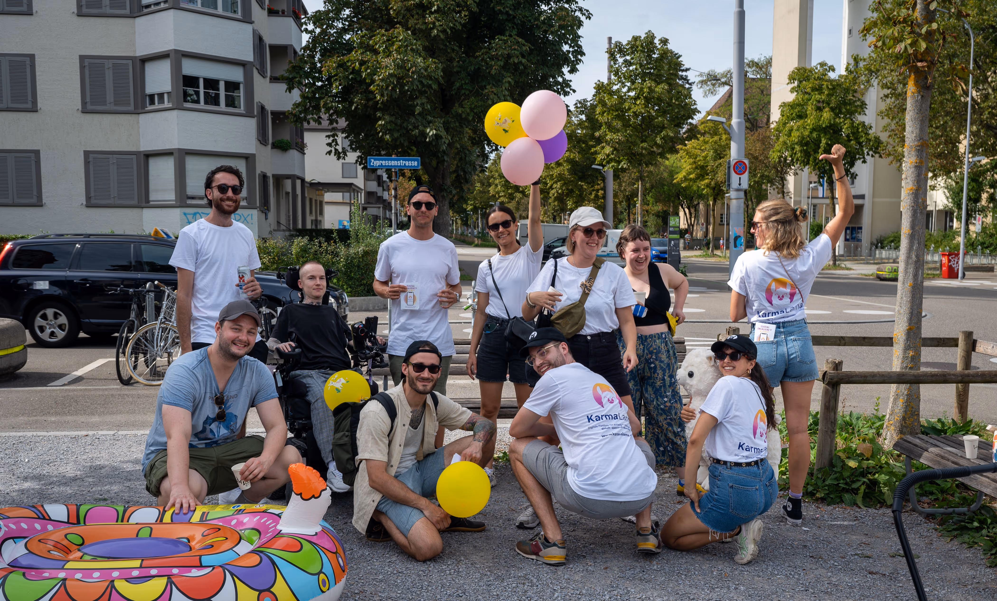 Gruppe junger Menschen, einige mit weißen 'KarmaLama' T-Shirts, posieren mit bunten Luftballons und einem aufblasbaren Lama im Freien an einer Straßenecke.