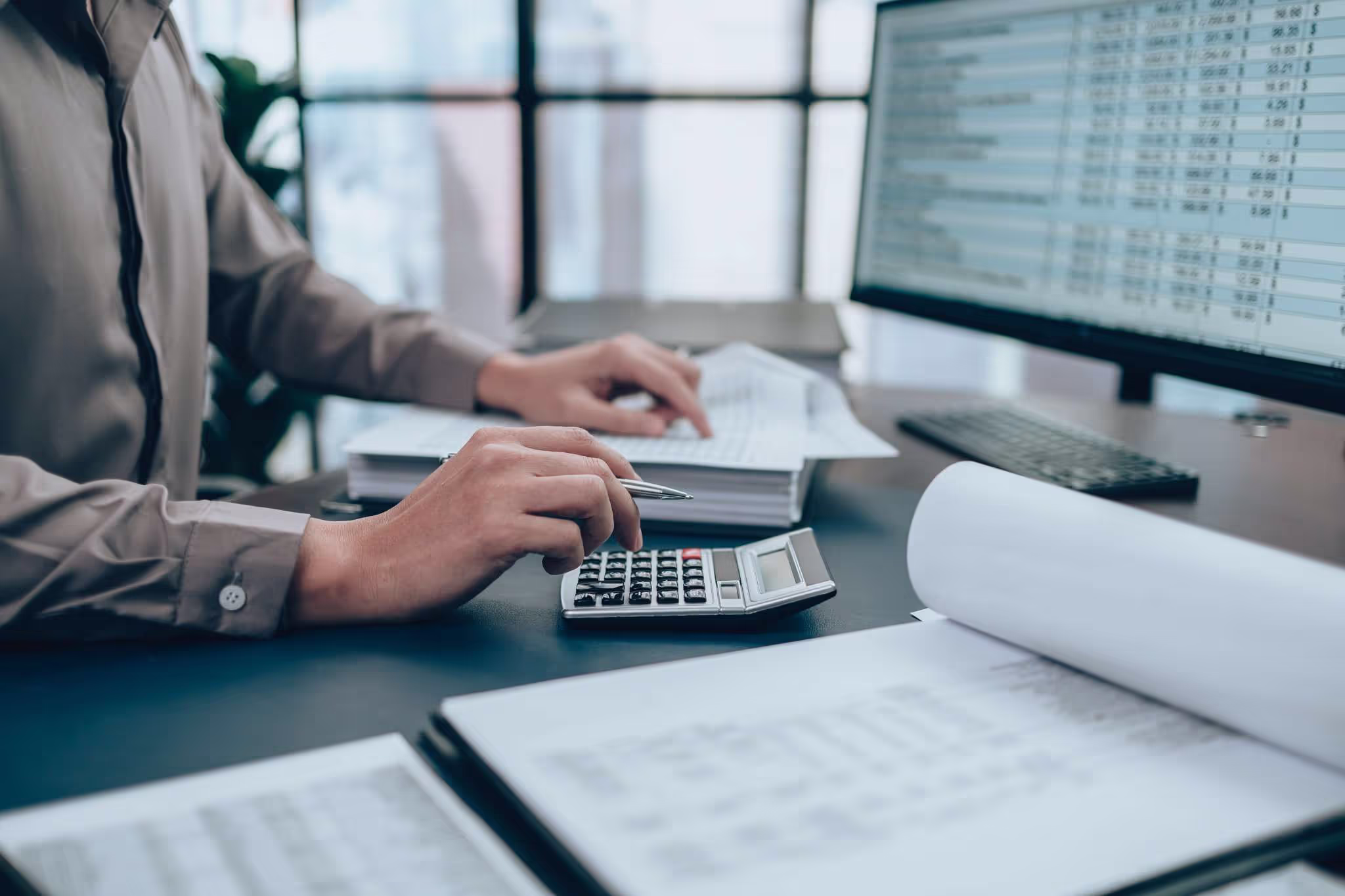 Person working at a desk with financial documents, a calculator, and a computer screen displaying spreadsheet data in an office environment