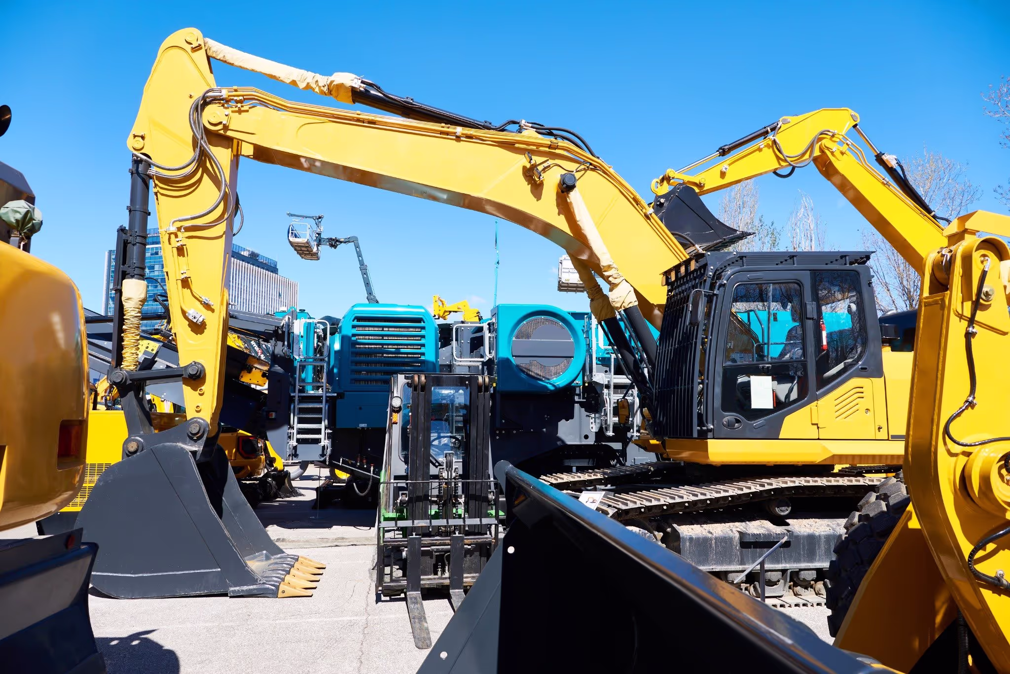 Heavy construction equipment including yellow excavators and industrial machines displayed outdoors on a sunny day, with hydraulic arms and attachments visible