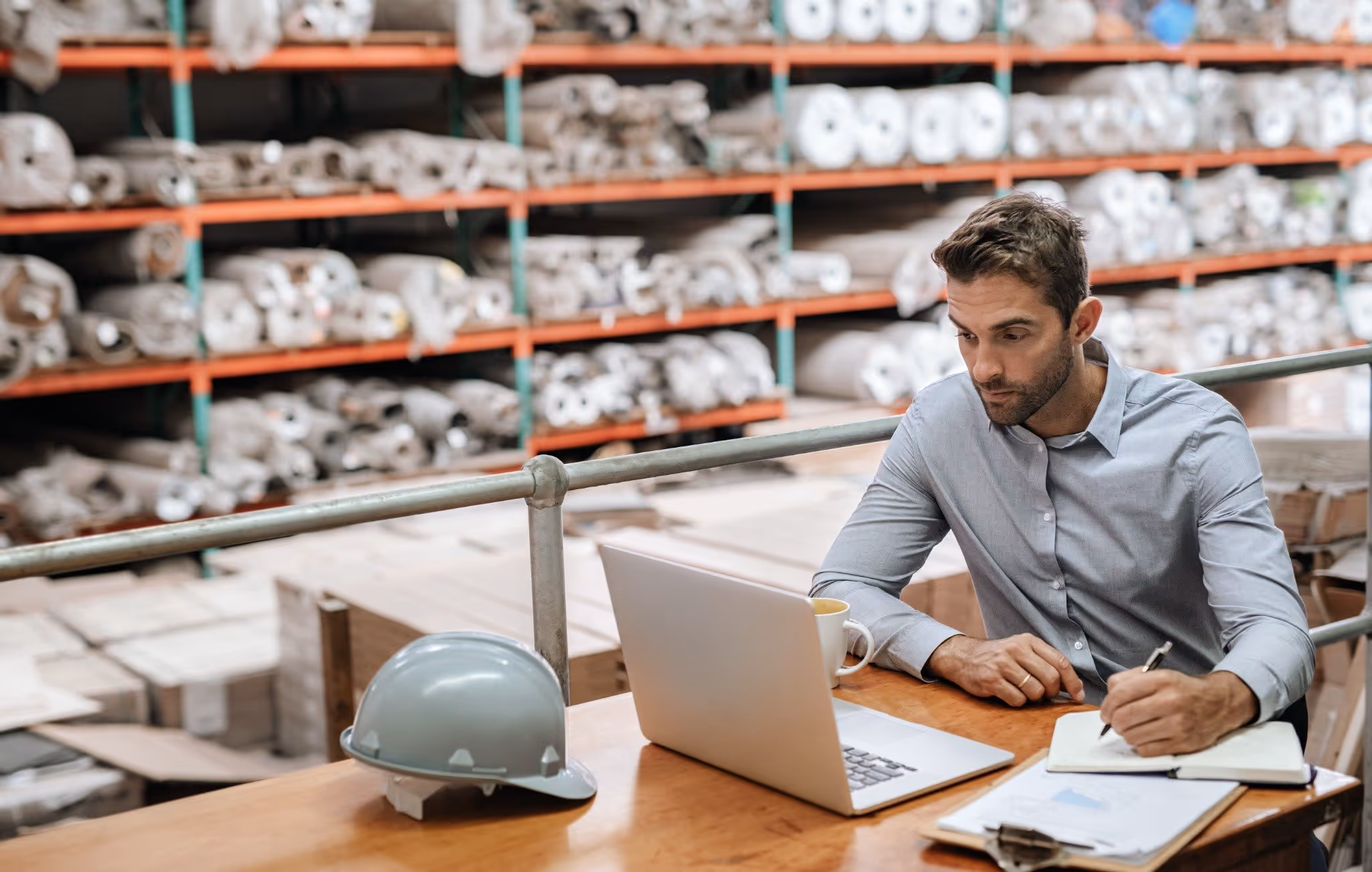 Man in suit using laptop and writing in notebook at a desk in a warehouse
