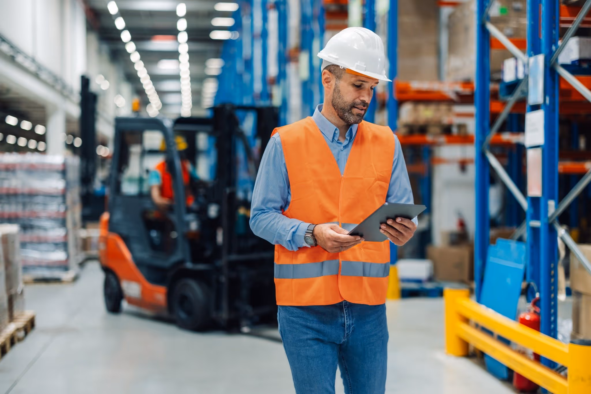 Warehouse worker in a hard hat and high-visibility vest using a digital tablet, with a forklift operating in the background among shelves and pallets