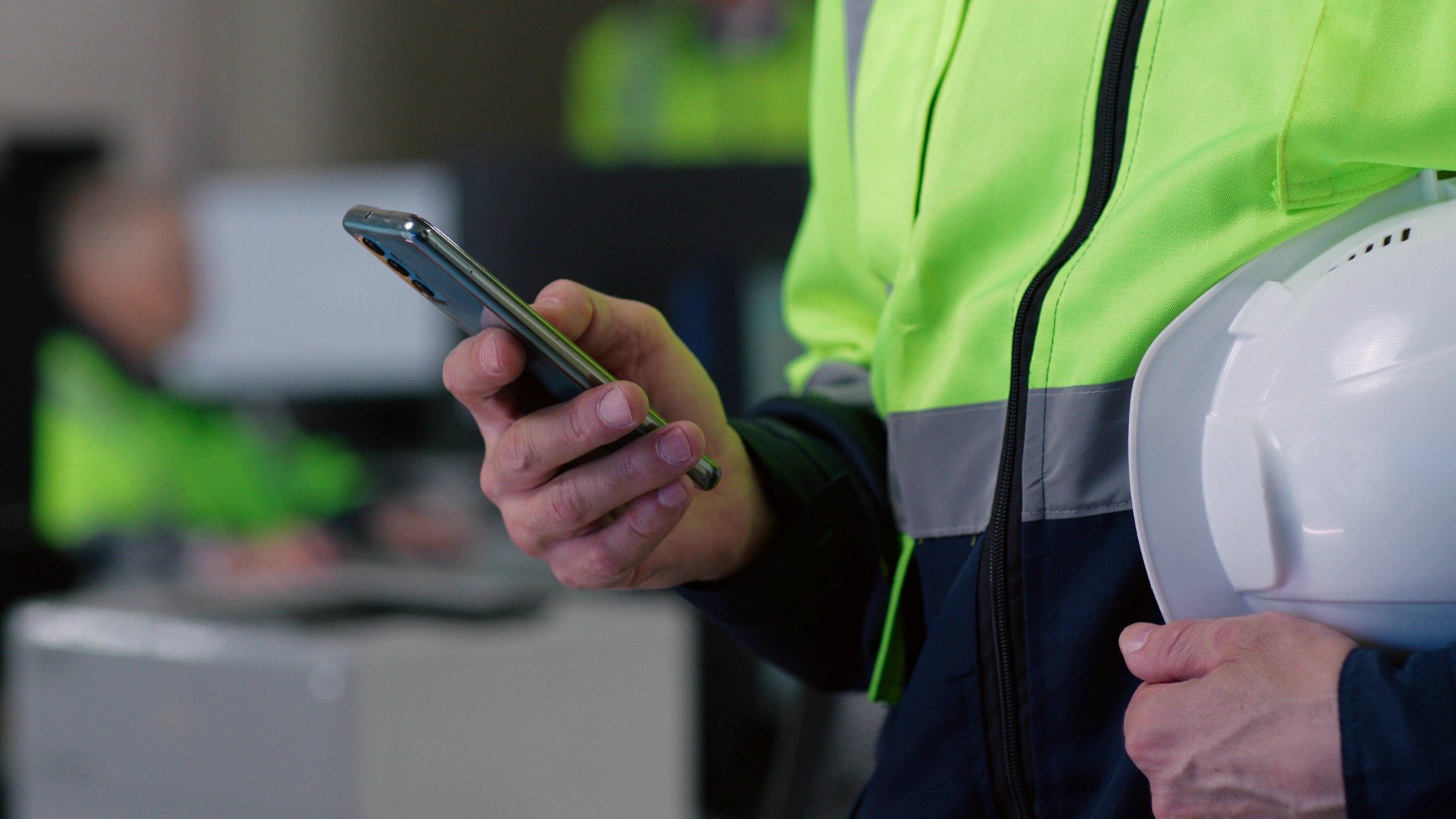 Close-up of a person in a high-visibility jacket holding a smartphone and a white hard hat