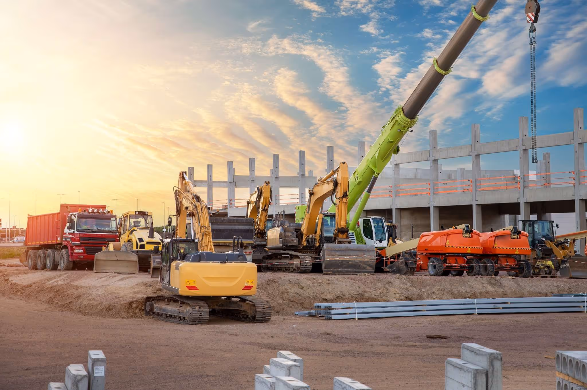 Construction site at sunset with various heavy machinery including excavators, a crane, dump truck, and rollers in front of a partially built structure