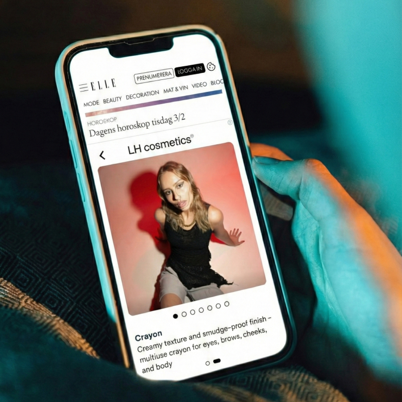 Hand holding a smartphone displaying a webpage about LH cosmetics featuring a woman posing and a description of a creamy, smudge-proof crayon for eyes, brows, cheeks, and body.