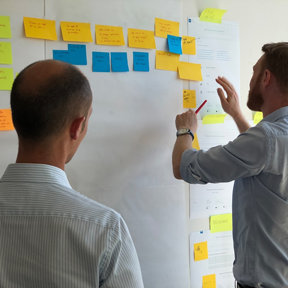 Zwei Männer arbeiten an einer Wand mit Notizzetteln und Dokumenten, während einer von ihnen mit einem Stift auf eine Idee zeigt. Symbolisiert den kreativen und strategischen Prozess hinter der Fallstudie für Hugo Boss.